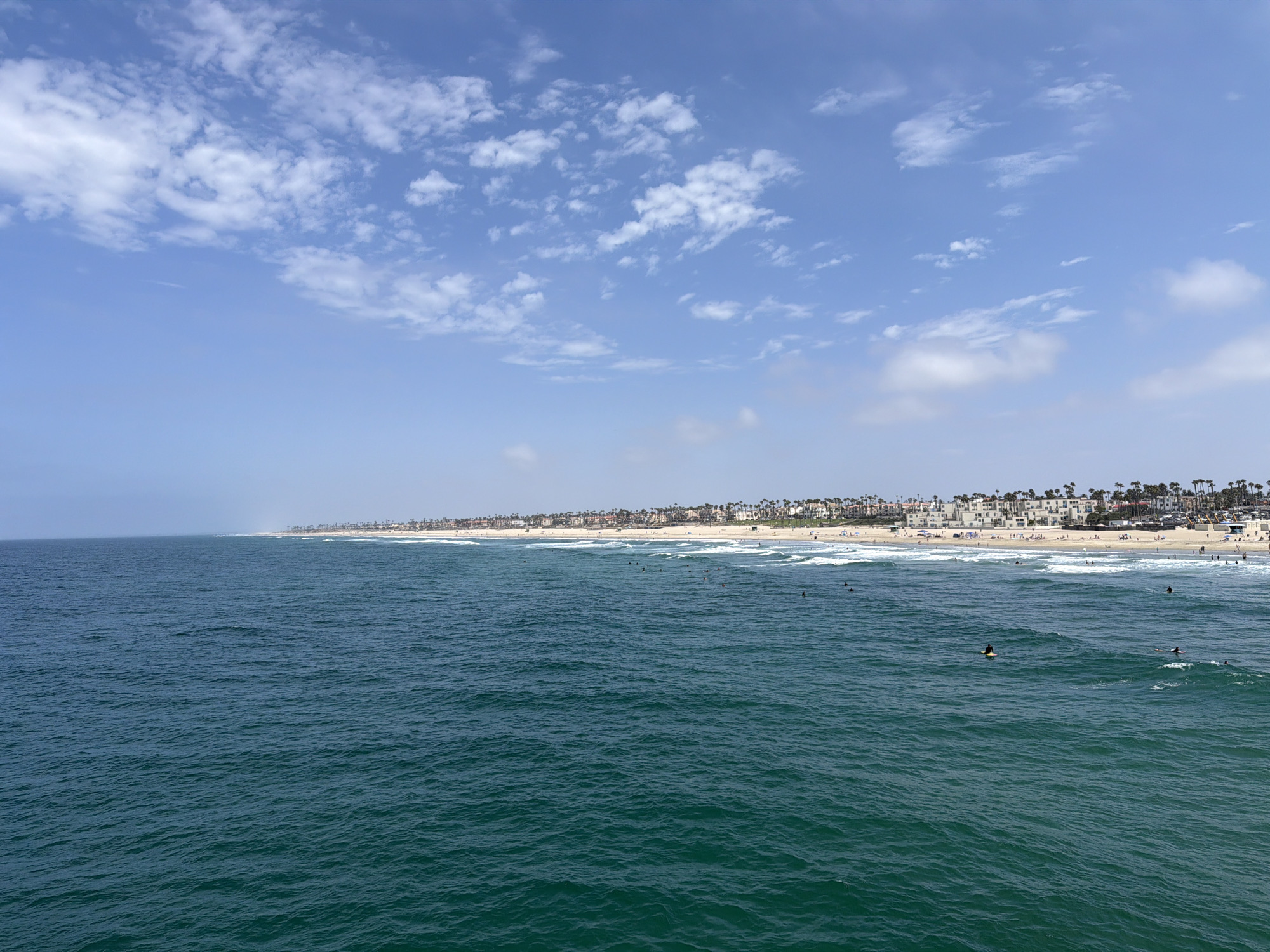 A wide-angle, eye-level, medium shot captures the vast expanse of the Pacific Ocean meeting the shore of Huntington Beach, California, on a clear, sunny day. The water, a deep teal hue, is slightly choppy with gentle waves rolling towards the sandy beach. In the foreground, several surfers are visible in the water, some on surfboards and one actively riding a wave. Beyond the surf, the shoreline is lined with a dense row of buildings, varying in height and architectural style, many with balconies and tiled roofs, suggesting a coastal city. Palm trees are interspersed among the buildings, and the beach itself is dotted with small figures, presumably beachgoers. The sky above is a brilliant blue, adorned with scattered white cumulus clouds, enhancing the sense of a perfect beach day. The image is static and depicts a serene, yet active, coastal scene.