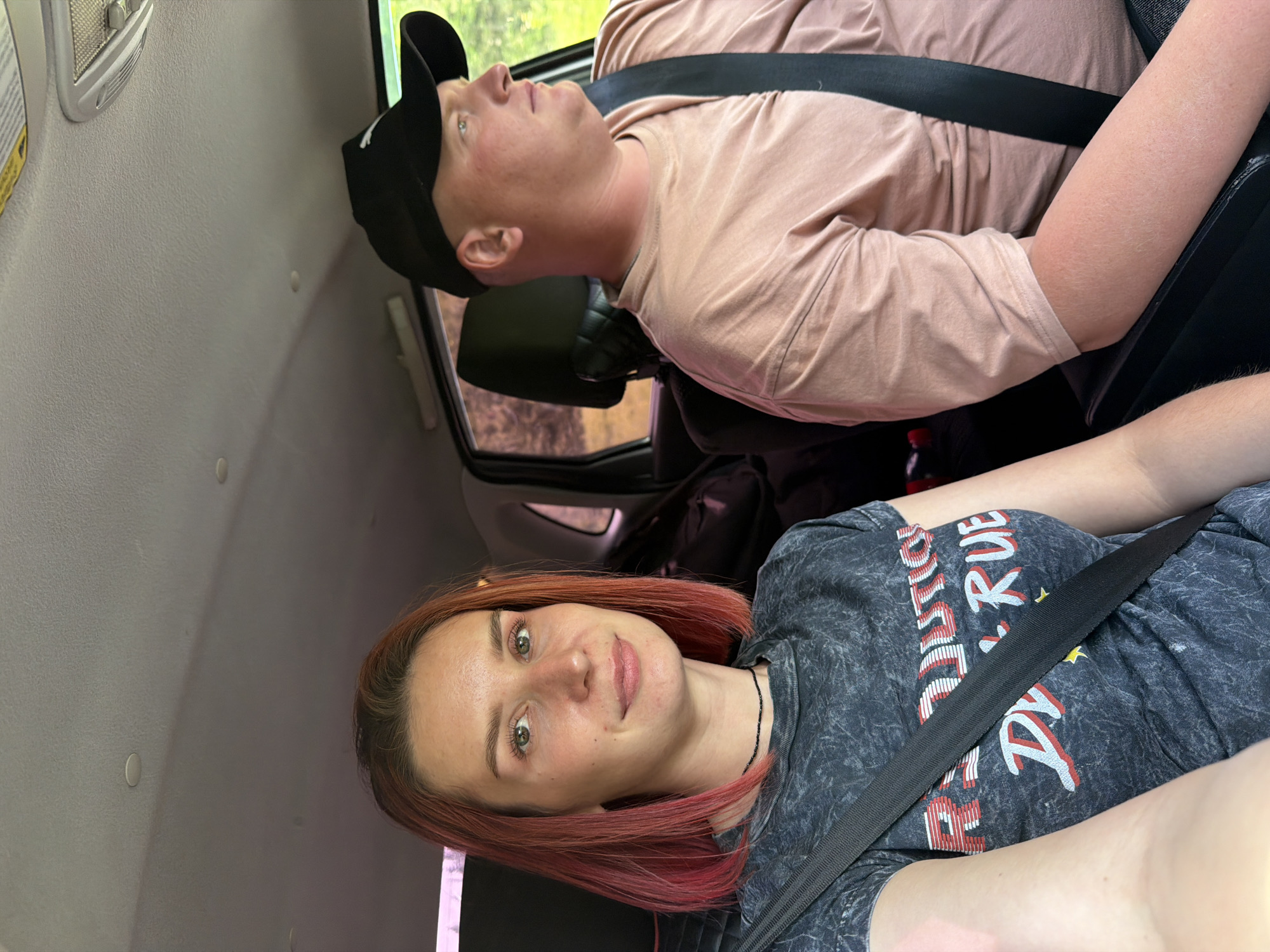 Two people are in a vehicle. The person in the front passenger seat is looking at the camera. This person has reddish-brown hair and is wearing a gray t-shirt with a design. The person in the back is looking up and toward the driver's side window. This person is wearing a black baseball cap and a pink t-shirt. Both are wearing seatbelts. The interior of the vehicle is light gray. The location is possibly the city of Горняк, Russia, but this cannot be confirmed from the image alone. The environment appears to be daytime.