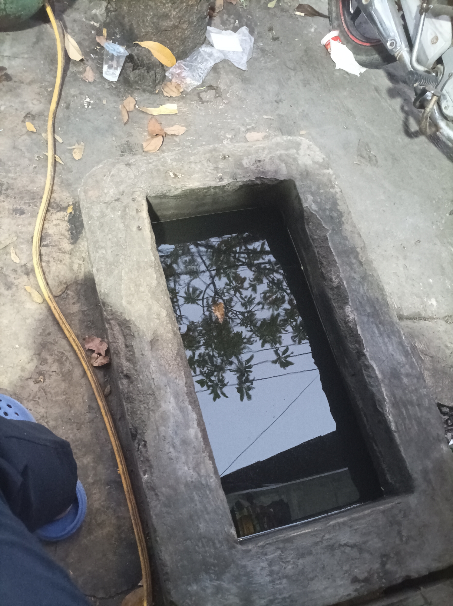 The image shows a rectangular concrete pit filled with dark water, reflecting tree branches and a building. The pit is situated on a cracked concrete surface that appears to be an outdoor area, possibly a yard or alleyway, in Cirebon, Indonesia. Scattered on the ground around the pit are dry leaves, a discarded plastic cup, and some white plastic debris. A yellow hose runs along the left side of the pit, and a person's legs, clad in dark pants and wearing a blue sandal, are visible in the lower left corner of the frame. In the background, parts of a motorcycle and its exhaust pipe are visible, suggesting the location might be near a road or a parking area. The overall impression is one of a somewhat neglected outdoor space with evidence of recent activity or debris accumulation.