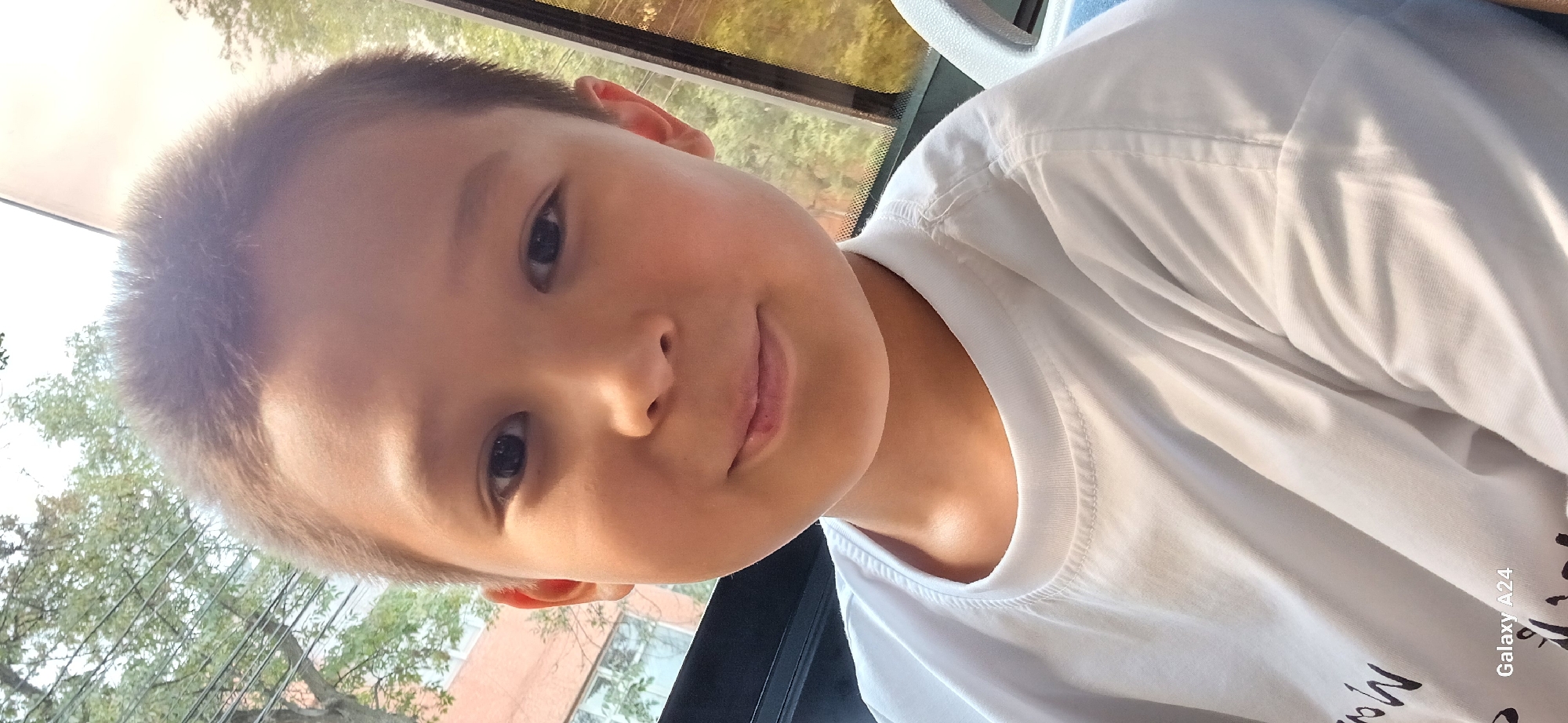 A young boy, likely under ten years old, is pictured in a close-up selfie. He has short, dark hair and is wearing a white t-shirt. His face is looking directly at the camera, and he has a slight smile. There is a small mark on his upper lip. The background suggests he is in a vehicle, possibly a bus or car, with a window on the right side showing trees and buildings outside. The bottom left corner of the image displays "Galaxy A24", indicating the device used to take the photo. The overall lighting is bright, suggesting it is daytime. The scene does not provide specific indicators to pinpoint a location within Bishkek City, Kyrgyzstan.