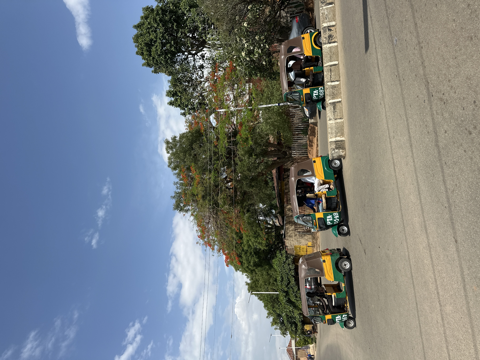 The image shows a street scene in Jos, Nigeria. Three auto-rickshaws are parked along the roadside, facing in the same direction. Each is a yellow and green vehicle with a brown roof and displays the inscription "PTA" followed by a number. Each auto-rickshaw contains a person. Trees line the side of the road, some with red flowers. The sky is blue with scattered white clouds. The road surface is gray asphalt. A sidewalk with a curb is adjacent to the vehicles. A gray car is parked beyond the auto-rickshaws, further along the street. Power lines are visible above the street. Buildings are visible in the background.