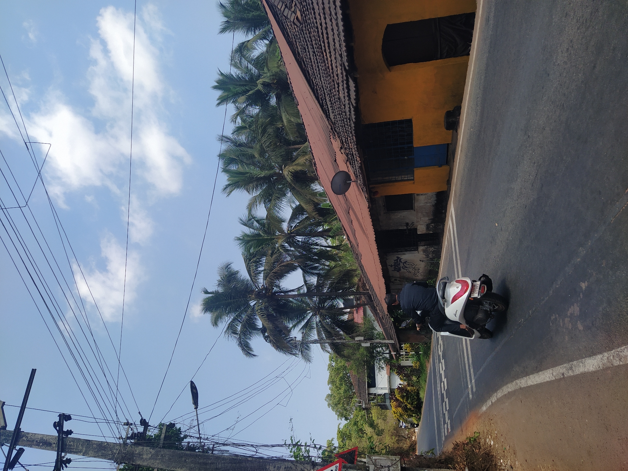 A street scene in Moira, India shows a man on a white scooter parked at the side of a road. The scooter is positioned next to a yellow building with a tiled roof. Several palm trees tower above the building. Overhead power lines stretch across a blue sky with some clouds. Other vegetation and buildings are visible in the background. The road has white lane markings.
