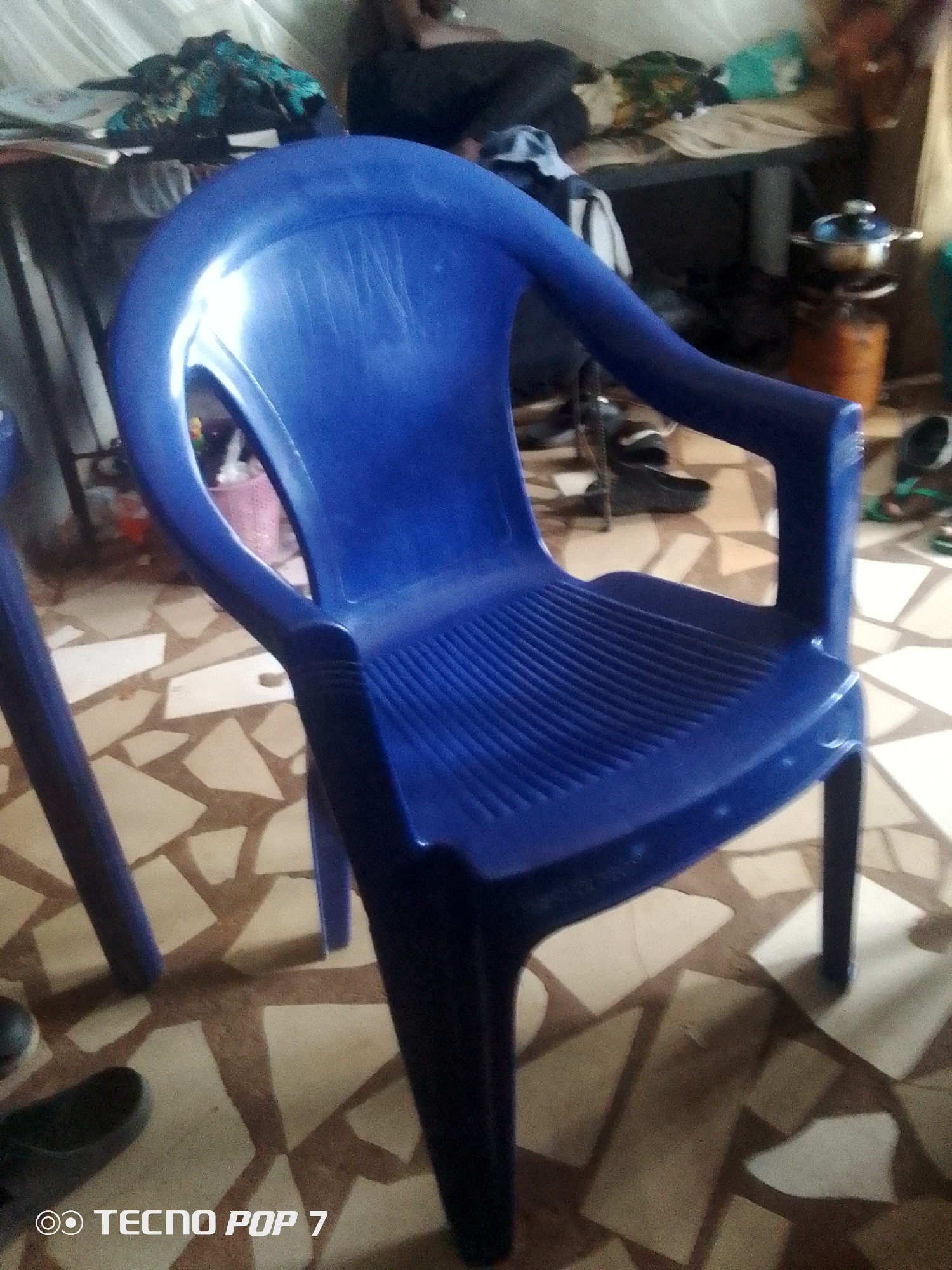 The image shows a close-up of a blue plastic chair in a room. The chair is the primary subject, with its ribbed seat and back clearly visible. The floor beneath the chair appears to be tiled with irregular geometric shapes in shades of beige and brown. In the background, out of focus, there are hints of other objects, including what looks like a table with some fabric draped over it, and a metallic pot on a burner. The lighting suggests an indoor environment, possibly with natural light coming from the side. The presence of the "TECNO POP 7" watermark at the bottom suggests this is a photograph taken with a Tecno Pop 7 mobile phone. Without further context, the exact location within Ipetumodu, Nigeria, cannot be definitively determined from the image alone. The scene appears to be a casual domestic or informal setting.