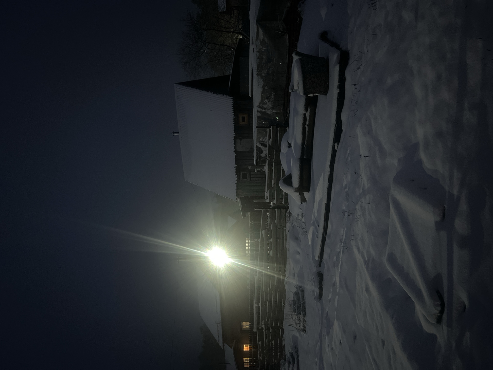 The image shows a snow-covered landscape in Chaikovsky, Russia, at night. A building with a snow-covered roof is visible, along with a fence. A bright light source illuminates the scene, creating a starburst effect. The foreground is covered in snow, and a dark sky is visible in the background. Some windows are lit in a house.
