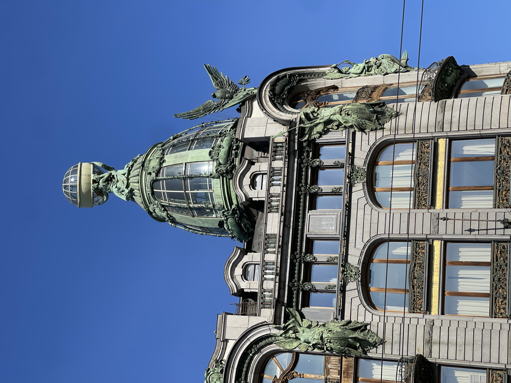 The image shows a portion of the Singer House, an Art Nouveau building in Saint Petersburg, Russia. The structure is predominantly gray with bronze-colored ornamentation. The most prominent feature is a glass dome topped with a sphere. Above the dome, a large bronze eagle with outstretched wings perches. The building's facade includes large, arched windows with ornate metal detailing. Bronze sculptures of figures are positioned above and around the windows and dome. The sky is a clear, bright blue.