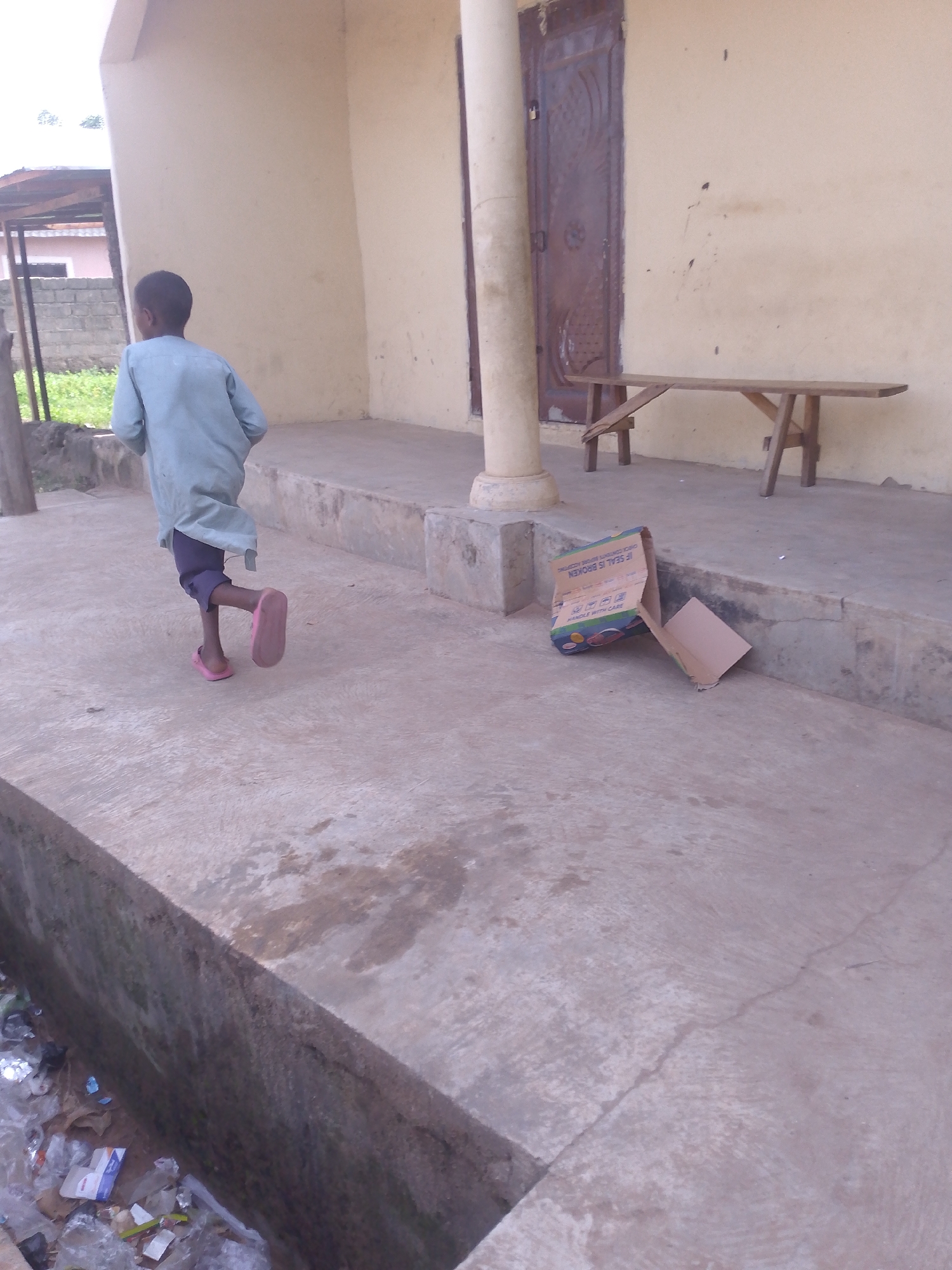 In Serti, Nigeria, a young boy wearing a light blue tunic and dark trousers is captured mid-stride, his right leg extended as he runs towards the right of the frame. He is barefoot, with pink sandals on his feet. The background reveals an outdoor setting with a building and a wooden bench. The building's exterior wall is a pale yellow, marked with dark splatters, and features a large, dark, ornate door. A thick, cylindrical pillar stands to the left of the door. In the foreground, a concrete walkway is visible, sloping down and to the left, where accumulated debris, including plastic bags and paper packaging, lies in a lower area. To the right of the boy, a cardboard box, partially open and with printed text, rests on the edge of a raised concrete platform, with a flap extended onto the walkway. A simple wooden bench, supported by A-frame legs, is positioned against the building wall. The overall scene suggests an outdoor area adjacent to a structure, possibly residential or commercial.