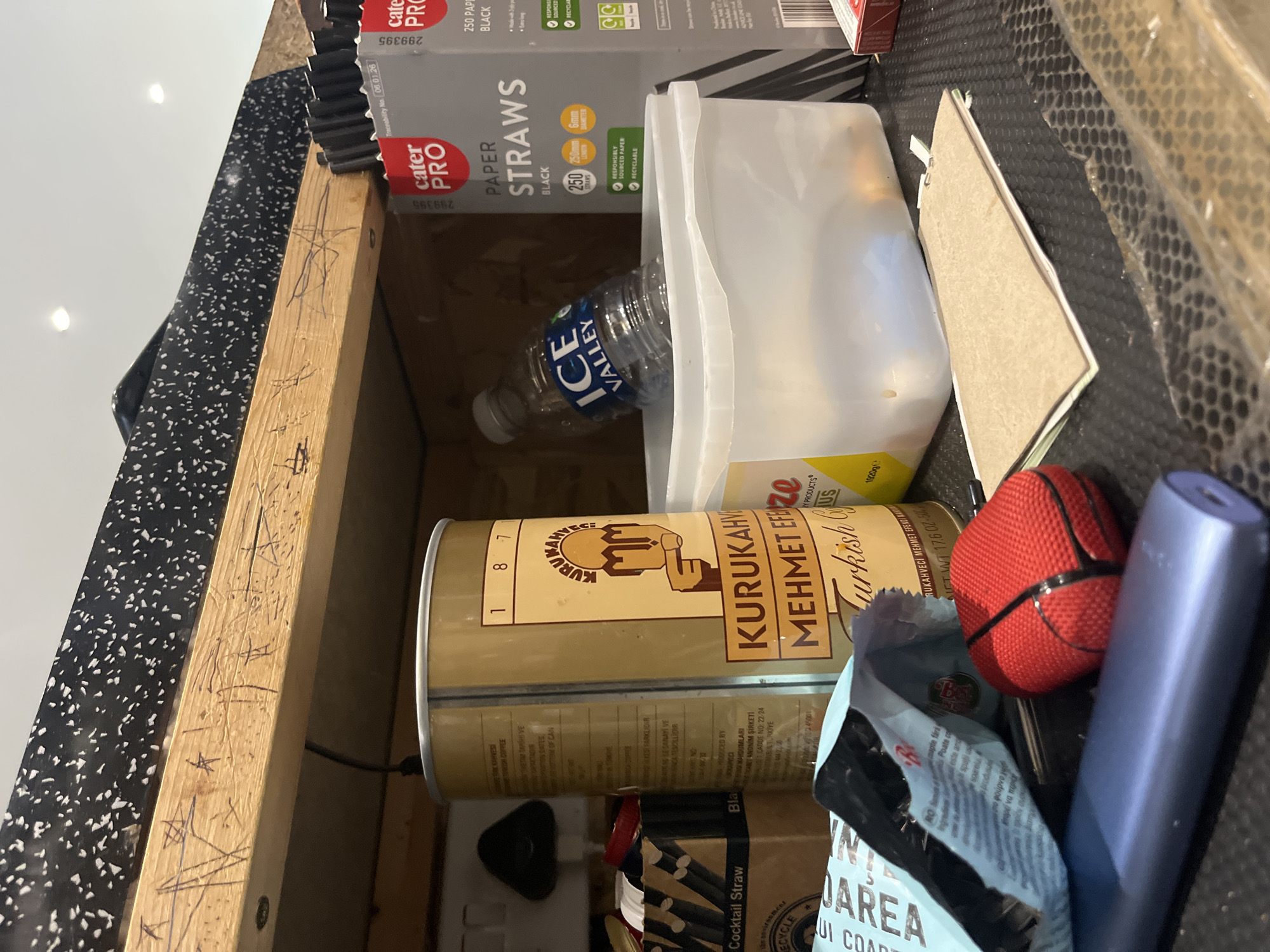 A cluttered storage space, possibly a drawer or cabinet, contains various items. A cylindrical tin of "Kurukahveci Mehmet Efendi Turkish Coffee" dominates the lower portion of the image. Beside it is a blue package with "UI COAREA" partially visible. A red, round object (perhaps a ball) and a slim, blue device are adjacent. Above the coffee tin is a clear plastic container, partly obscuring a "Ice Valley" water bottle. "Cater Pro Paper Straws" (black) are visible in their packaging. There is a piece of cardboard among the items. The cabinet or drawer is constructed of light wood, and the lining is a black material with white speckles. The location is not discernible from the image.