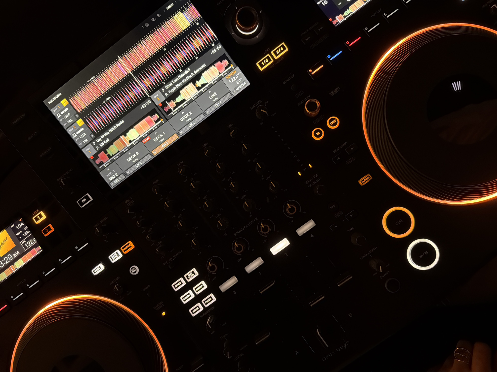 The image shows a close-up view of a Pioneer Opus Quad DJ controller in a low-lit environment. The controller features various illuminated buttons, knobs, and a central screen displaying waveforms and track information. The screen shows the names of two songs being played: "Day 'N' Nite (WILO Remix)" by Kid Cudi and "Ghost Town (Extended)" by Purple Disco Machine ft. Retrosonix. Different controls such as "Beat FX", "Sound Color FX" and "Master Tempo" are identifiable. Part of a hand with a silver ring can be seen in the lower right corner of the image. No further detail regarding a location in Budva, Montenegro, are present.