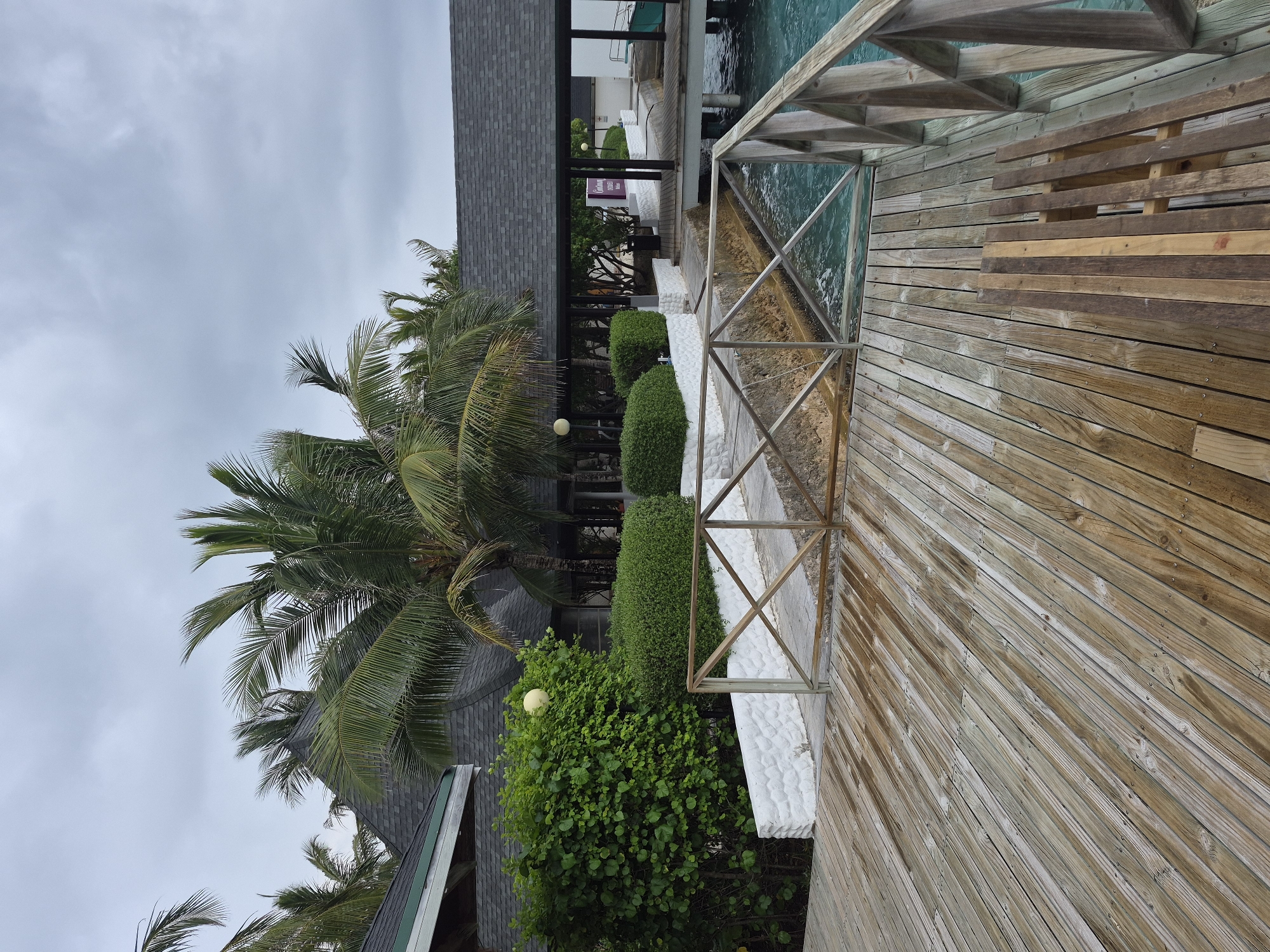 The image shows a wooden deck overlooking the ocean in Huraa, Maldives. The deck has a metal railing and a set of wooden stairs leading to the water. There are several buildings in the background with gray shingled roofs, partially obscured by lush green vegetation including palm trees and sculpted hedges. The sky is overcast. The boardwalk appears wet, indicating recent rain or high humidity.
