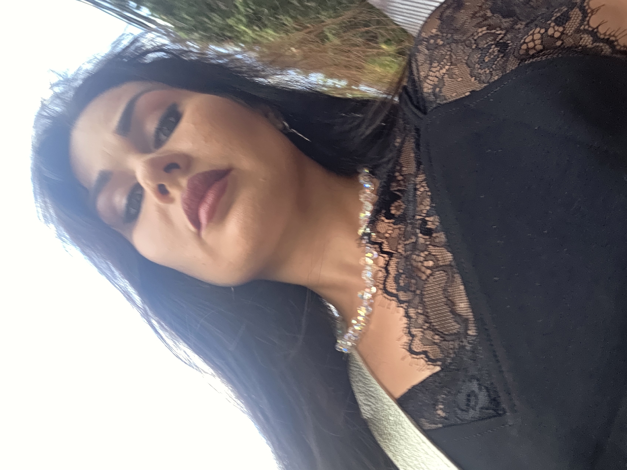 A close-up, low-angle, outdoor selfie features a woman with dark hair and makeup. She is wearing a black top with lace detailing on the shoulders and neckline, and a crystal necklace. Her face is the primary focus, with her lips slightly parted and her eyes looking into the camera. The background shows greenery and a bright sky, suggesting an urban environment. There are no explicit indicators pointing to a specific city like Sirius, Russia, within the image itself.