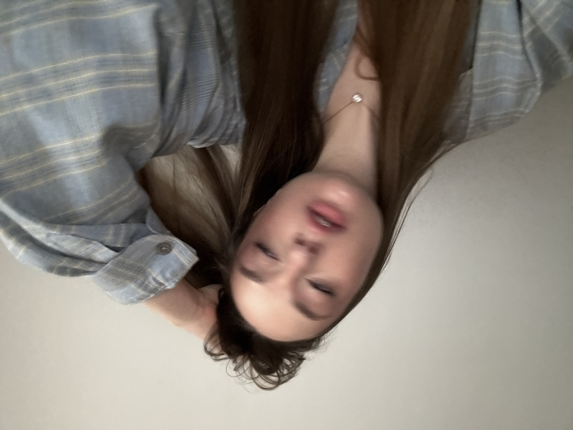 A young woman is lying down, facing the camera, wearing a light blue and white striped shirt and a delicate necklace. Her hair is brown and long. The background is a plain, light-colored wall.