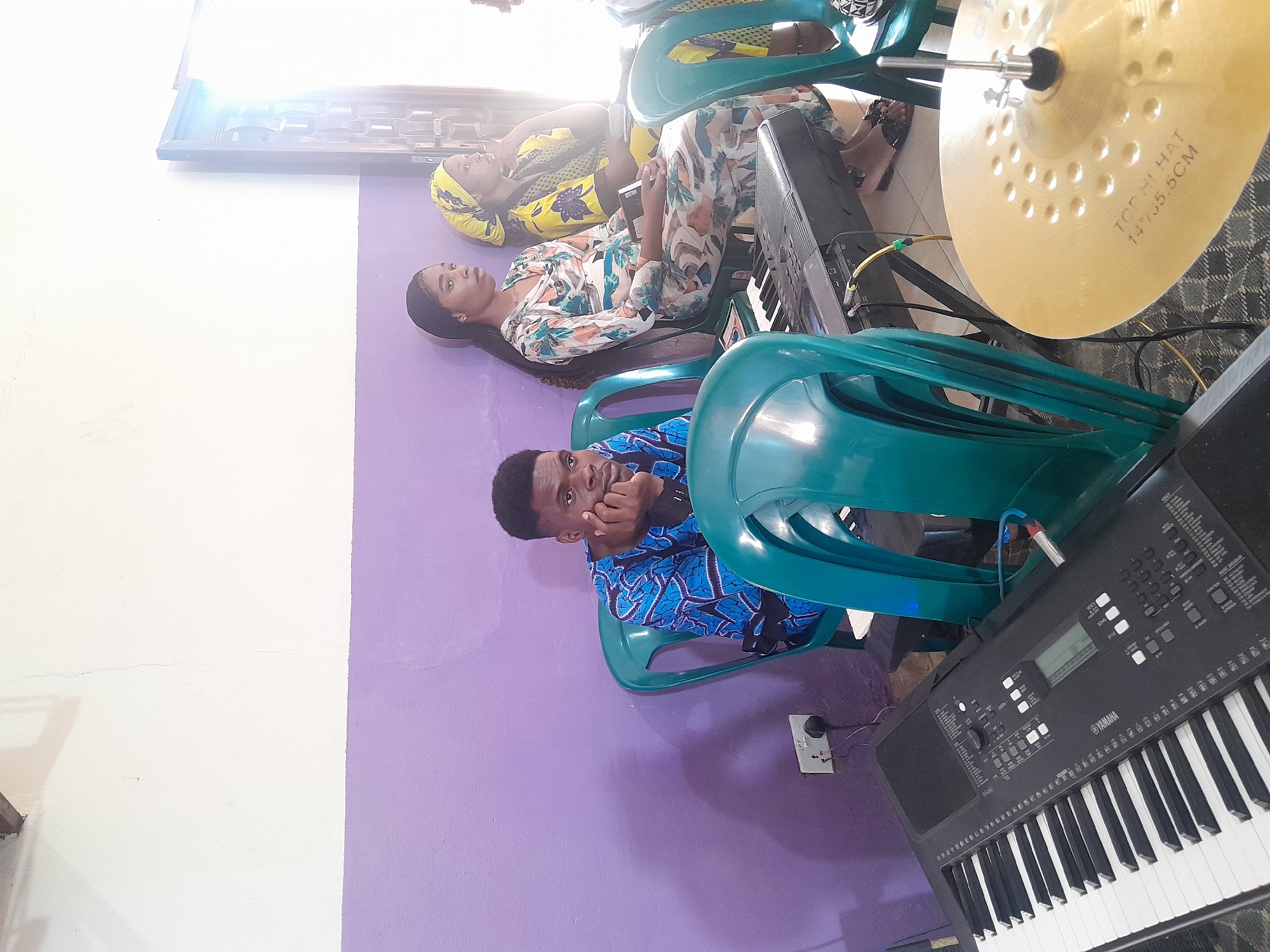 The image shows an indoor scene in a room with a wall that is half white and half lavender. Three individuals are sitting on green plastic chairs.
The person closest to the camera is a dark-skinned man wearing a blue patterned shirt. He is looking directly at the camera with his hand to his face.
Behind him and slightly to the left is a woman with dark hair, wearing a floral print dress. She appears to be looking upwards.
Behind the woman is another woman wearing a yellow headscarf and a patterned top. She also seems to be looking upwards.
To the right of the man is a black Yamaha keyboard. A gold-colored cymbal is positioned above the keyboard. An electrical outlet is visible on the lavender wall near the man's chair.
The image does not provide enough information to determine the specific location within Jos, Nigeria.