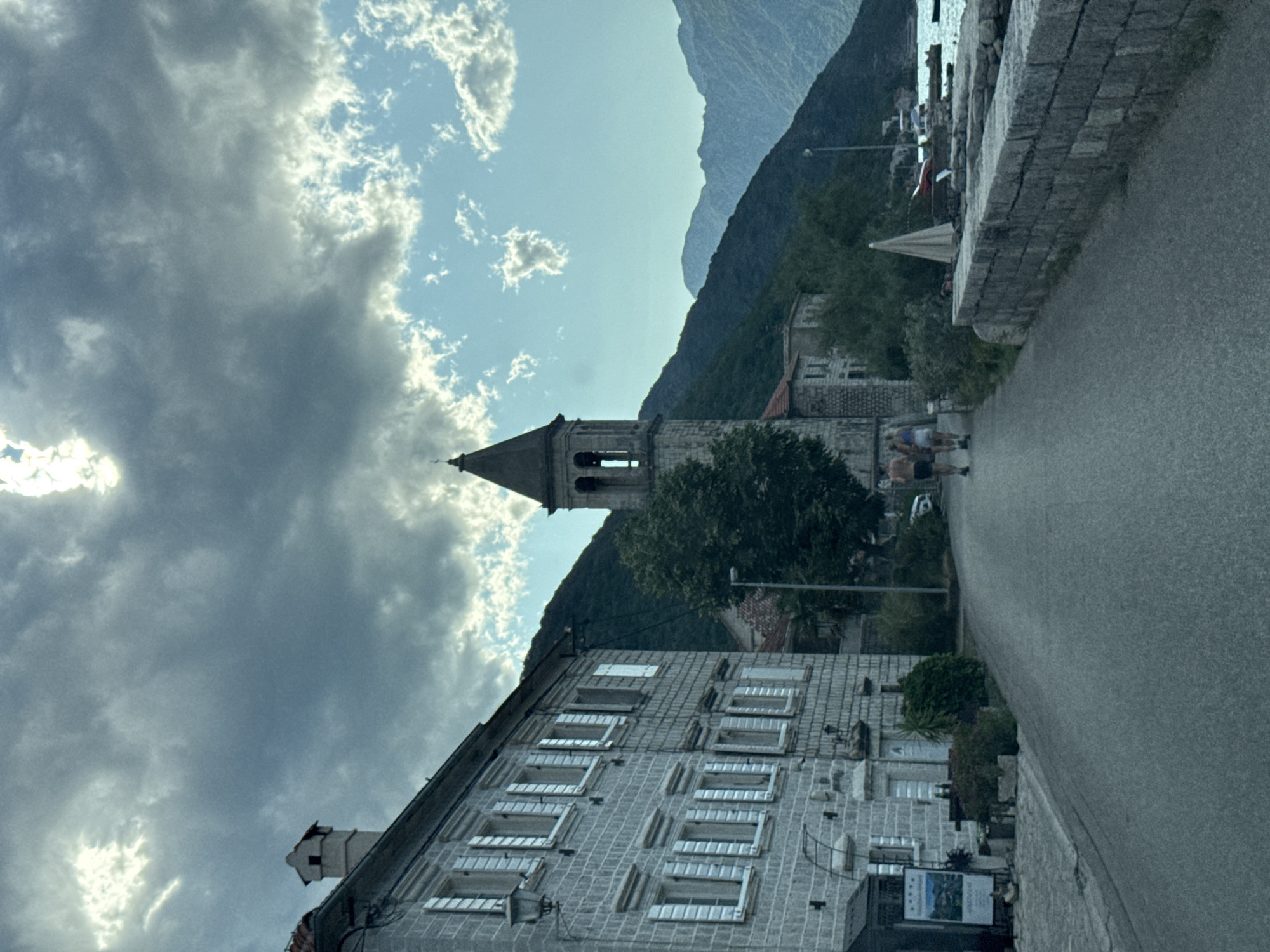 A view down a paved road in the coastal town of Prčanj, Montenegro, with a stone building on the left and the sea and mountains on the right. The sky is partly cloudy, with dramatic light breaking through. The building on the left is made of stone with white shutters. A bell tower is visible further down the road, and mountains rise behind the town. Two figures are walking away from the camera on the road. A stone wall lines the right side of the road, overlooking the water where several boats are moored.