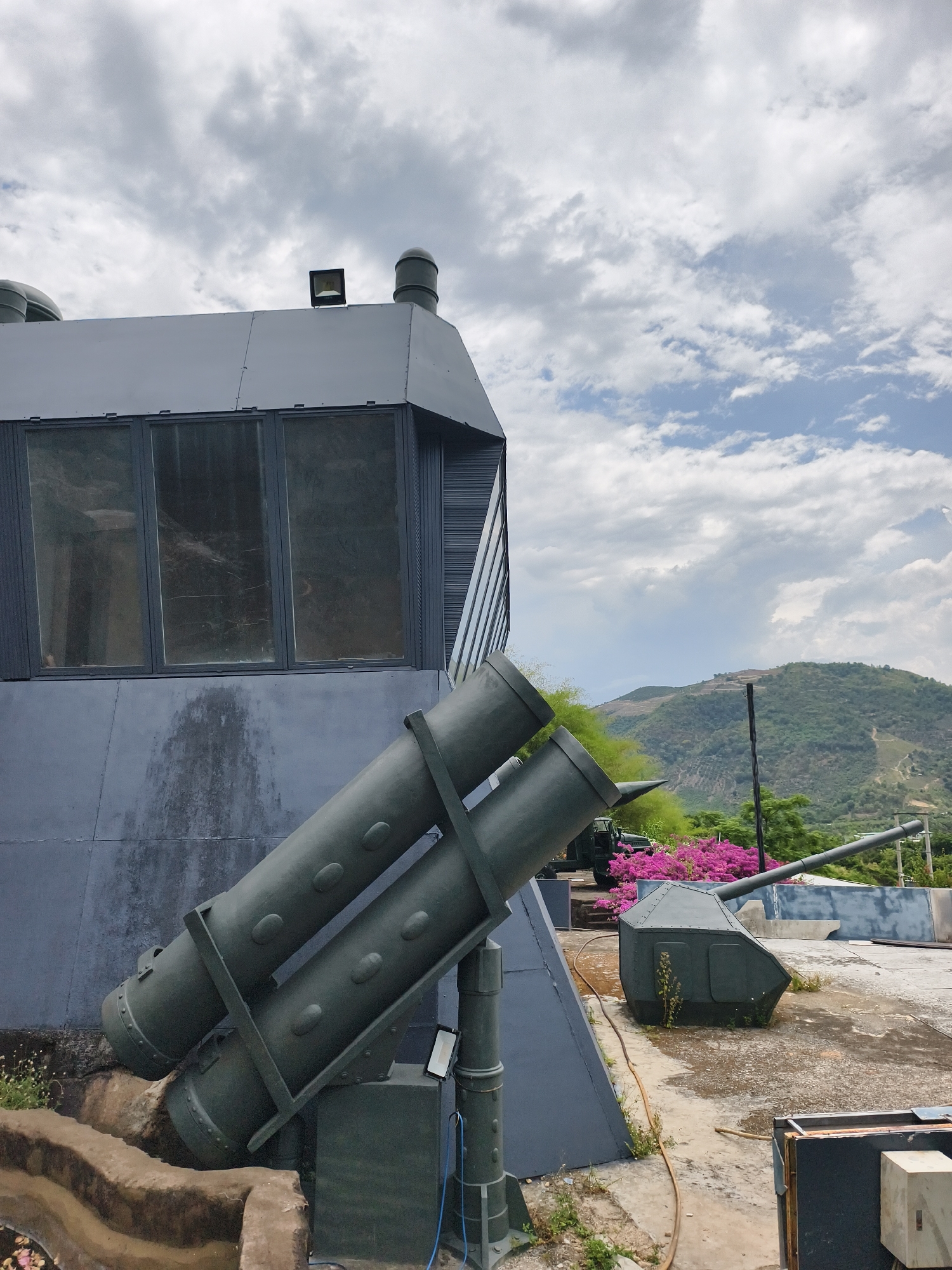 The image depicts a gray building with military-themed installations, located in Tay Nha Trang Ward, Vietnam. A two-story section with windows tops the building, which has a flat roof. In the foreground, there are two sets of parallel, cylindrical structures resembling missile launchers. Behind this, a gun turret sits on the roof. The background includes a mountainous landscape under a sky with white and gray clouds. Pink bougainvillea bushes are visible near the turret. The roof is concrete and shows some weathering.