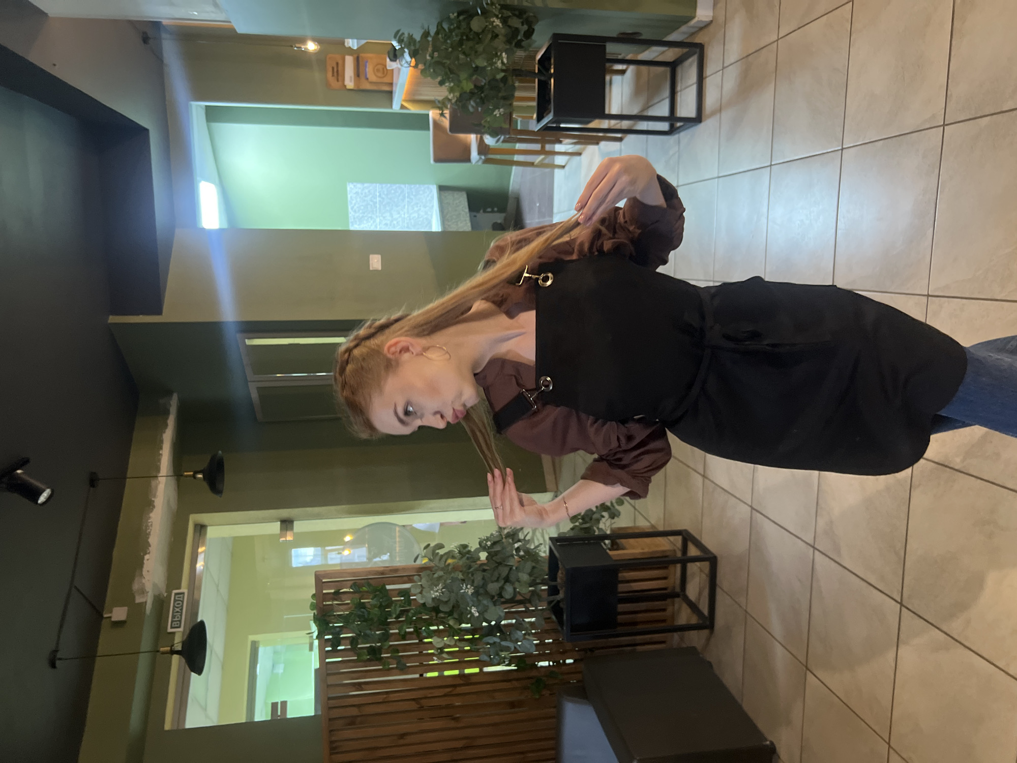 A full-length indoor shot depicts a young adult female posing playfully within a casual eatery, likely a cafe or restaurant, located in Красные Баки, Russia. The woman stands centrally, facing slightly left while looking right, pouting her lips and holding a strand of her light brown hair in each hand. She wears a long-sleeved dark brown blouse, a black full apron with two prominent brass-colored strap rings over the chest, and blue jeans. Accessories include small gold hoop earrings and a thin bracelet on her right wrist.
The environment features olive green walls and a dark grey or black ceiling with exposed track lighting and two black pendant lights suspended over a doorway on the left. Above this doorway, a white sign with black text reads "ВЫХОД" (EXIT), and a patch of unpainted or unfinished white drywall is visible near the ceiling. The floor is covered in light beige square tiles with dark grey grout lines.
To the immediate left of the woman, a dark grey upholstered armchair or bench is partially visible. Further left and behind her, a tall wooden slat partition stands next to a black metal planter holding a medium-sized artificial plant with green leaves and small white flowers. To the right, another identical plant in a black metal planter is situated. In the background, a light-colored bar with a wooden slatted facade and two brown upholstered bar stools is visible. Stacks of what appear to be menus or paper containers are on the bar counter. Through an archway in the background, a room with lighter green walls and a section of tiled wall is discernible, lit by an overhead fluorescent fixture.