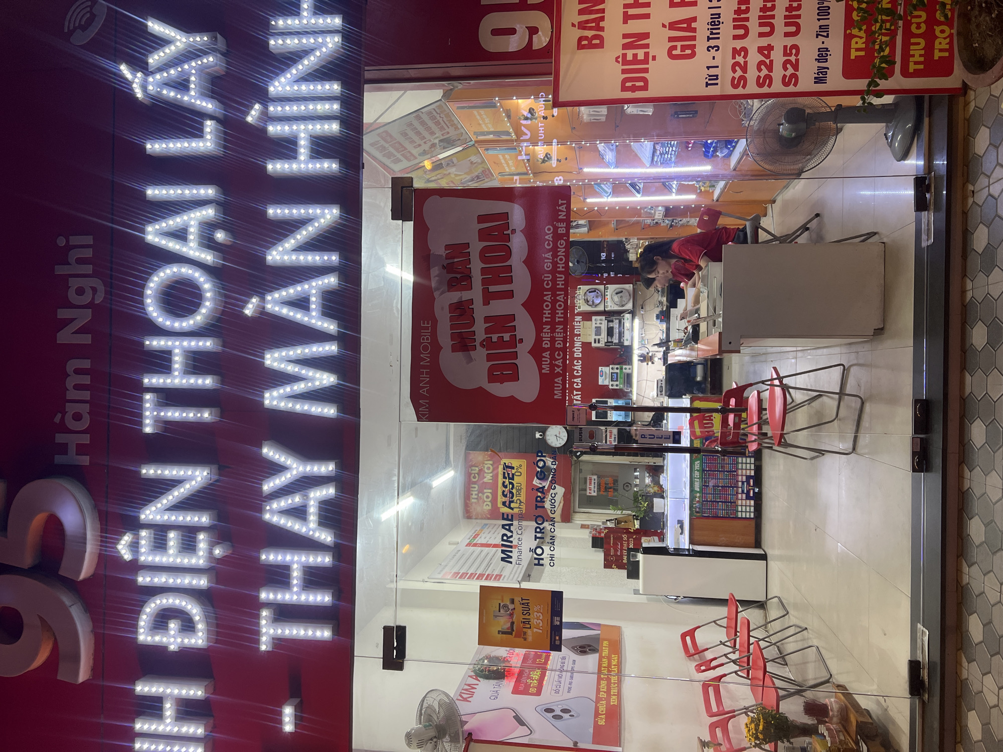 This is a photograph taken from outside a store, likely a mobile phone shop, in Da Nang, Vietnam. The store's facade is dominated by a large, bright LED sign that spells out "ĐIỆN THOẠI LẤY THAY MÀN HÌNH," which translates to "Phone Exchange Screen." The sign is illuminated with white LED lights, creating a striking visual effect, especially in what appears to be nighttime.
Inside the store, visible through the glass doors and windows, there are shelves displaying various electronic devices, likely phones and accessories. A young woman wearing a red shirt is seen working inside the store, interacting with equipment. Several red chairs are arranged outside the store, suggesting a waiting area or customer seating.
Several promotional posters are displayed on the store's exterior. One poster advertises "MUA BÁN ĐIỆN THOẠI" (Buy and Sell Phones), another offers "THU CŨ ĐỔI MỚI" (Trade-in Old for New) with "MIRAE ASSET Finance Company" and mentions a 0% interest rate. Another sign promotes "HỖ TRỢ TRẢ GÓP" (Installment Support) with the requirement of a citizen ID card. Prices for phones are indicated as ranging from 1 to 3 million Vietnamese Dong, with specific models like S23, S24, and S25 Ultra mentioned. The phrase "Máy đẹp - Zin 100%" (Beautiful phone - 100% Original) suggests the quality of used phones.
The overall scene conveys a busy commercial environment, with the bright signage and promotions highlighting the services offered by the mobile phone store. The presence of the chairs and the work being done inside indicate active business operations.