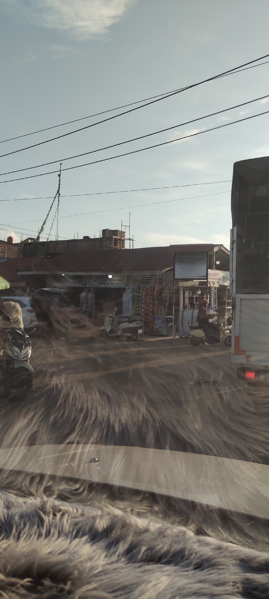 The image depicts a street scene in Meunasahmesjid, Indonesia. In the foreground, a car's windshield is covered with a fuzzy grey material, obscuring the view of the road ahead. The road surface itself is obscured by a dust or sand disturbance, creating a blurred effect that streaks horizontally across the image. On the left side of the road, a white motorcycle with license plate "BI 3655 GI" is parked. Further down the road, there are several shops with their fronts open to the street. One shop appears to be a "KARA MART". Scooters are parked in front of some of the shops, and people can be seen on them or walking. A large truck is parked on the right side of the road, its rear visible. Overhead, power lines crisscross the sky, and a vine-covered pole is visible. The sky is pale blue with some scattered clouds. The overall impression is of a busy street on a sunny day, with the dust disturbance adding a sense of movement or wind.