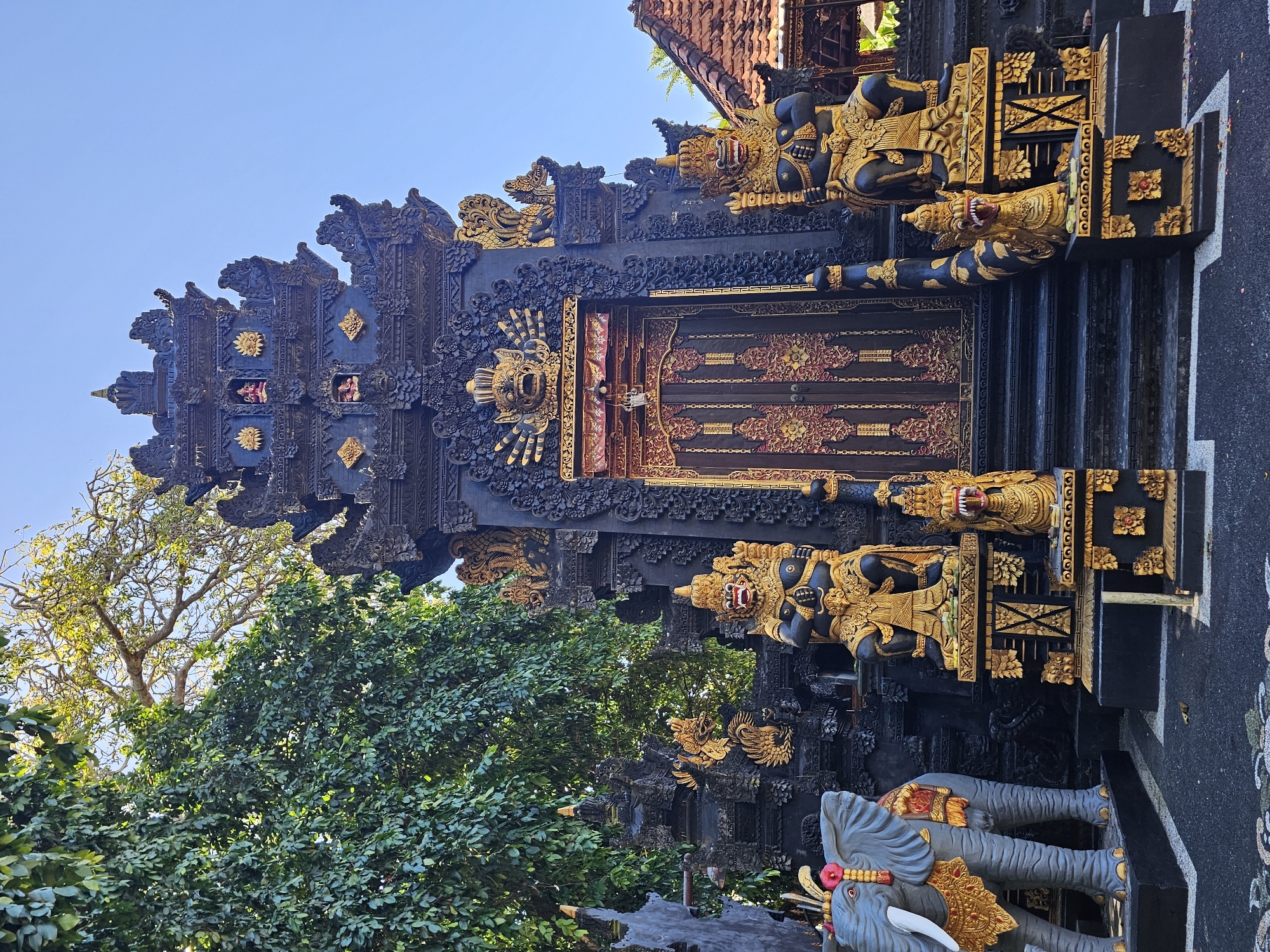 The image shows a traditional Balinese entrance gate, known as a "candi bentar," located in Kuta Selatan, Indonesia. The gate is predominantly black and gold, with intricate carvings and sculptures. Flanking the entrance are statues of deities and mythological creatures. At the base of the gate are two gray elephant sculptures. Lush green trees surround the structure, and the sky is clear blue.