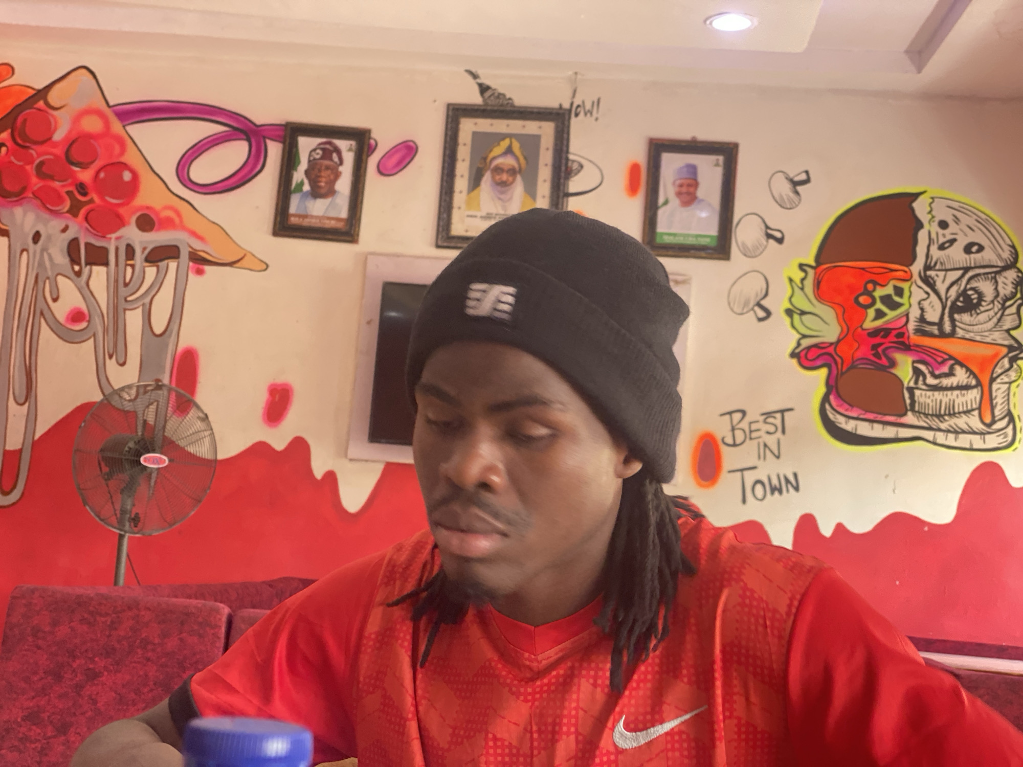 A man wearing a black beanie and a red shirt sits at a table in a restaurant in Tukur Tukur, Nigeria. Behind him is a wall decorated with food-themed graffiti, framed portraits of men, and a small television screen. An oscillating fan is also visible in the background. The restaurant wall is marked with the words "Best in Town".