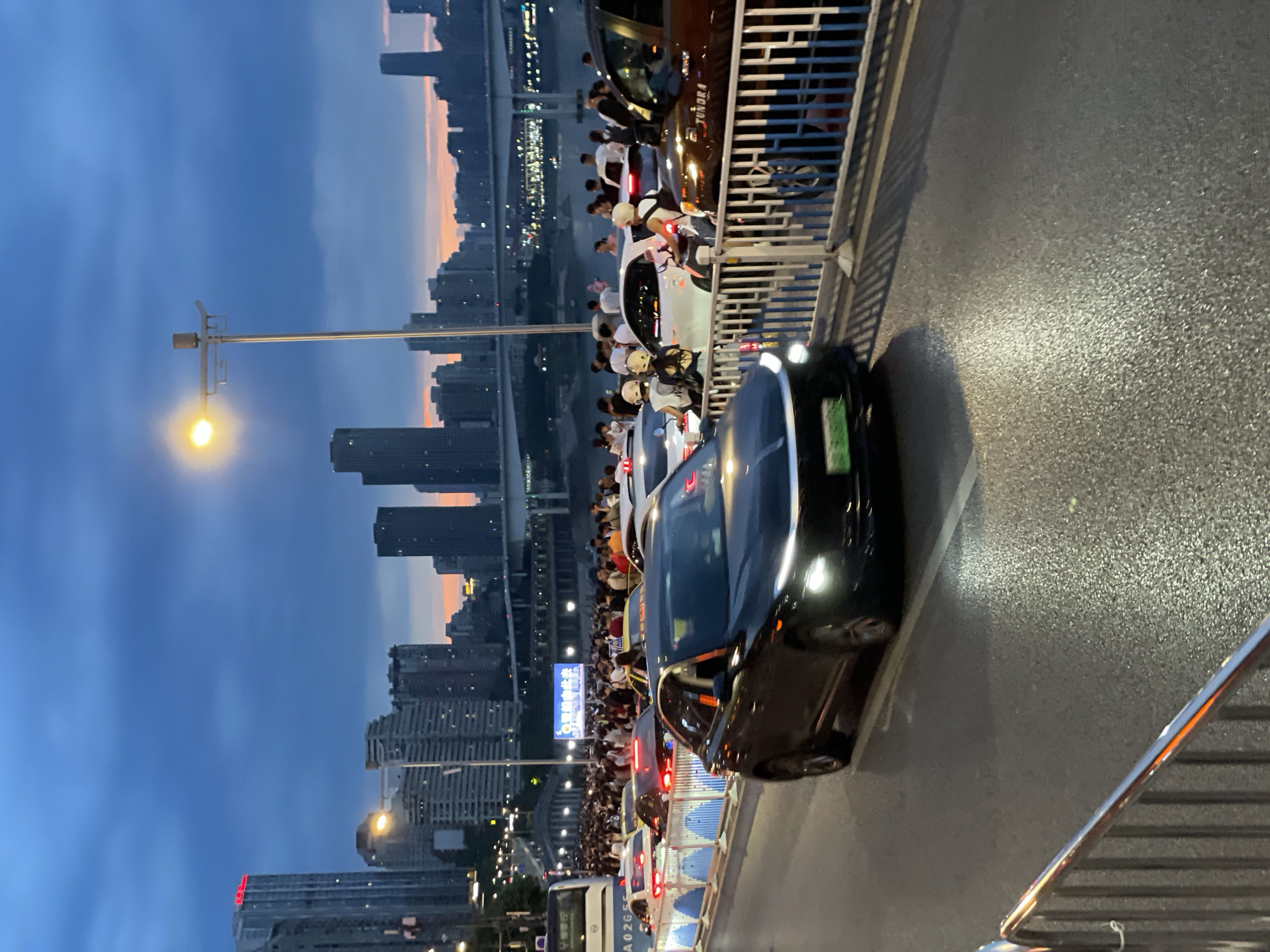 In Yuzhong District, China, at dusk, a congested street scene unfolds along a waterfront promenade overlooking a city skyline. The sky displays a gradient of deep blue to orange, indicating sunset. The foreground features a black car with its headlights on, driving on a wet asphalt road. A silver railing is visible in the lower left corner. Behind the car, a dense crowd of pedestrians lines a blue and white barrier, watching something off-camera. Several other cars, including a white sedan, a brown SUV, and a bus, are also present on the street. In the background, a river is visible, spanned by an elevated highway. The cityscape is dominated by tall buildings, illuminated by interior lights and the fading daylight. A streetlamp with a bright yellow light is positioned above the pedestrian area.