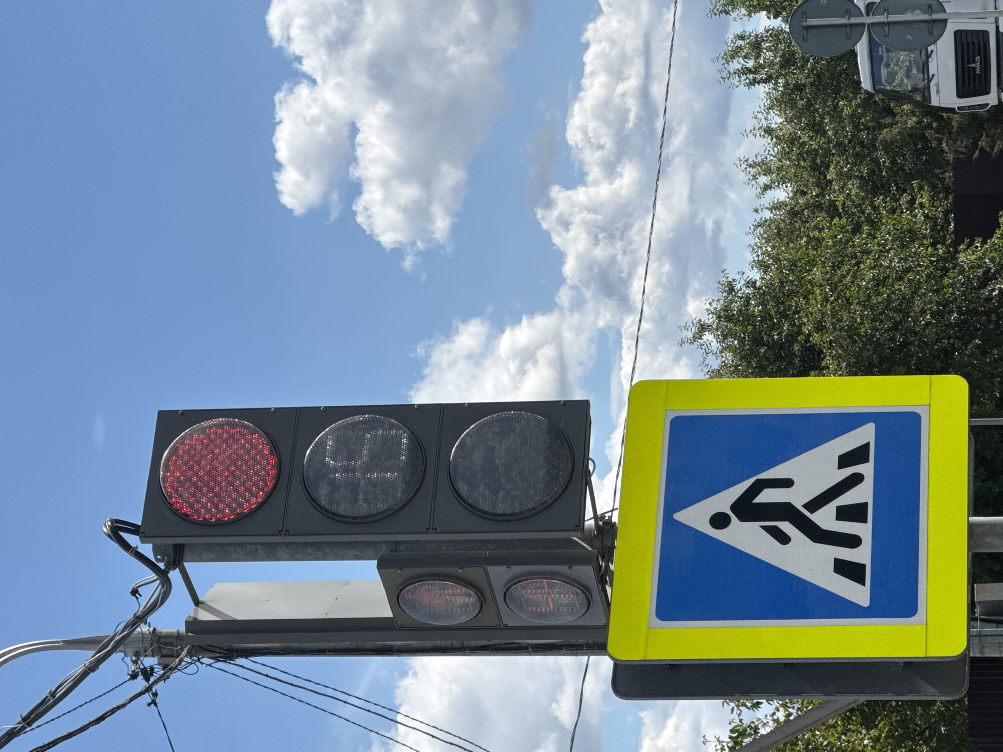 This media file shows a traffic light and a pedestrian crossing sign on a bright, sunny day. The traffic light has a red light illuminated, indicating that vehicles should stop. The pedestrian crossing sign is blue with a white triangle containing a black silhouette of a person walking across a crosswalk. The scene appears to be in an urban or suburban area, with trees and a portion of a white truck visible in the background. The sky is blue with scattered white clouds, suggesting good weather. The image is captured from a low angle, looking up towards the sky and the traffic control devices. There is no visible text in the media file. The overall impression is one of a typical street scene with traffic management in place.