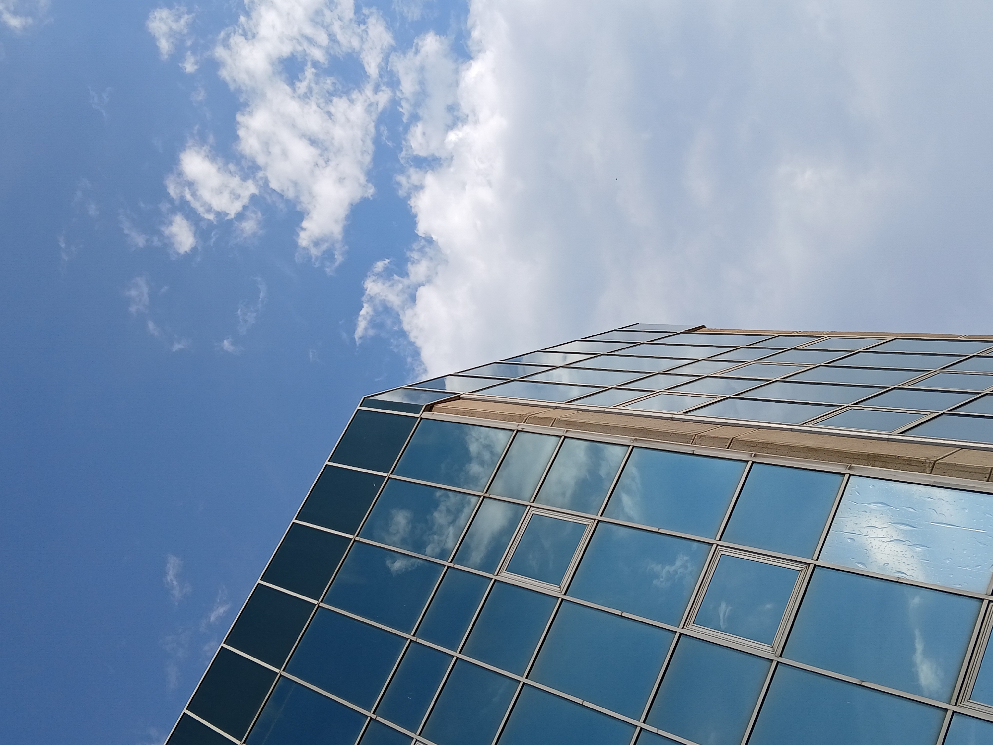 An architectural photograph captures a modern building with a reflective glass facade set against a dynamic sky. The building, angled diagonally from the lower left to the upper right of the frame, is composed of numerous rectangular and square glass panels framed by light-colored mullions. These panels act as mirrors, vividly reflecting the blue sky and scattered white clouds, creating an illusion of the sky extending into the structure. Some windows display faint streaks or droplets, possibly from rain or condensation.
The sky in the upper left is a clear, bright blue, transitioning to a lighter shade with varying densities of white, fluffy clouds across the rest of the upper portion of the image. The perspective is from a low angle, looking upwards, emphasizing the height and reflective quality of the building.
The scene depicts an outdoor setting during bright daytime, under sunny to partly cloudy weather. No individuals or specific activities are visible, presenting a serene, almost abstract view of urban architecture blending with nature. There is no visible text in the image. The geometric lines of the building and the organic shapes of the clouds create a striking contrast.