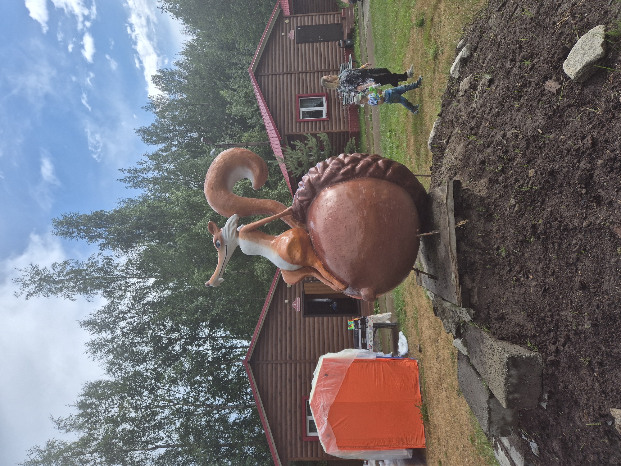 An outdoor scene depicts a large, whimsical sculpture of a squirrel sitting on an acorn, likely made of fiberglass or similar material, in a grassy area. The sculpture is situated in front of several rustic wooden cabins with red roofs, suggesting a resort or recreational area. Trees line the background under a partly cloudy sky.
To the left of the sculpture, an orange tent covered with a translucent plastic sheet is partially visible. A table with items on it, possibly for sale or display, is also present near the tent.
In the middle ground, two people are walking on the grass. A woman with blonde hair, wearing a patterned top and dark pants, is walking with a young boy dressed in a striped t-shirt and blue jeans, wearing a baseball cap. The boy appears to be holding a toy gun, possibly a water gun.
The ground in the foreground is a mound of dark soil with some scattered stones and sparse green shoots, indicating recent landscaping or unfinished construction. Concrete blocks are placed near the base of the sculpture.
The specific location is identified as Nizhnevartovsk, Russia. The context suggests a public or semi-public space, potentially a park, campground, or children's recreational facility. The overall atmosphere appears to be casual and family-oriented.