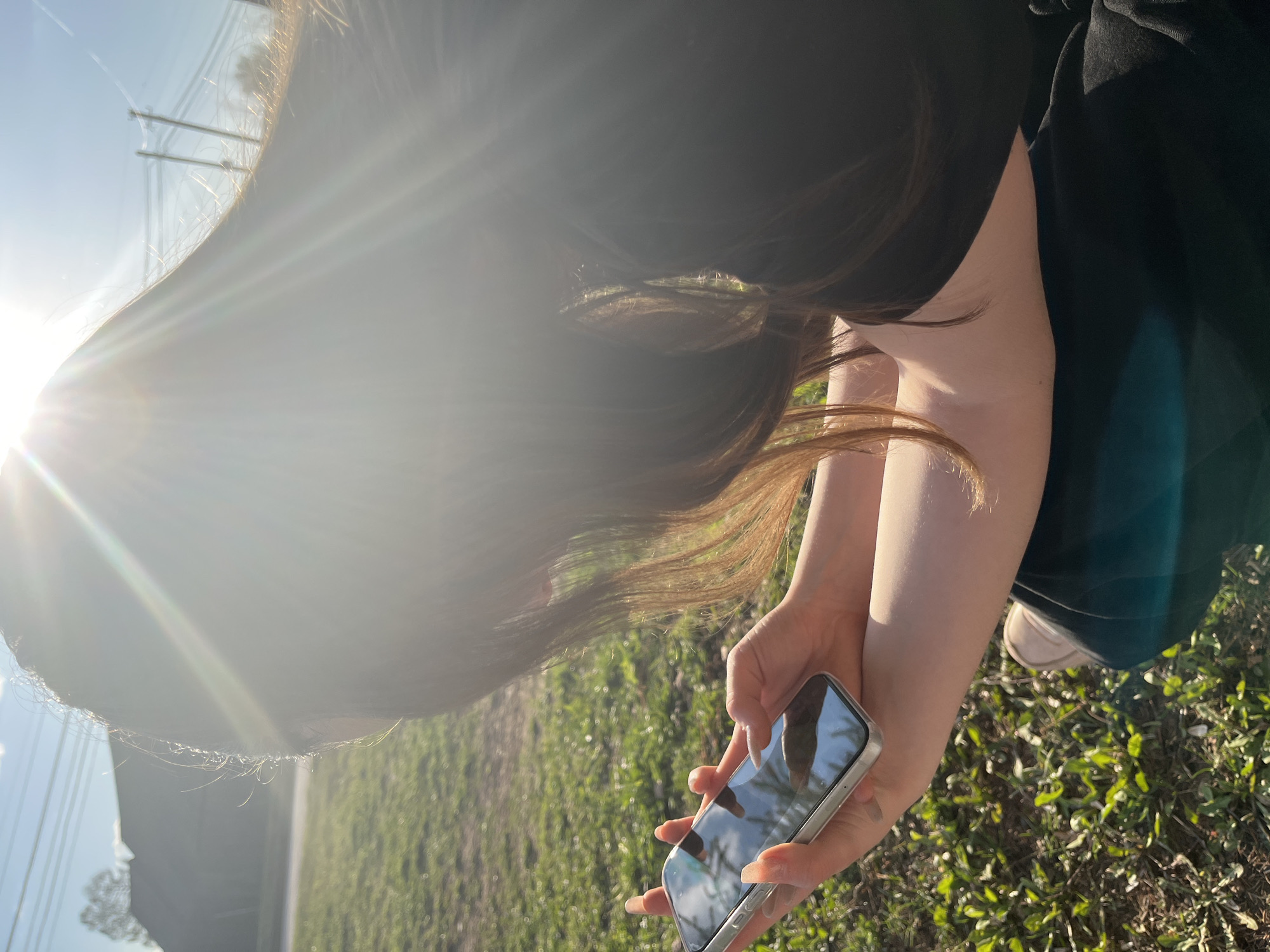 A female is standing outdoors in Сосново, Russia, using a smartphone. She is wearing a black top and a dark green skirt. Her long brown hair is falling forward. The grass is green, and a road and utility poles are in the background. The sun is shining brightly, creating a glare in the photo.