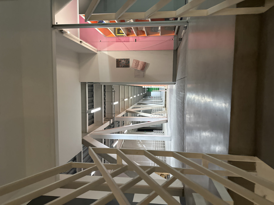 An upward-angled, straight-on perspective showcases a building's multi-story interior. White walls contrast with a gray concrete floor. A white, zigzagging stair railing spans multiple levels. Ceiling fixtures emit linear light. The floor above displays a vibrant, pink ceiling, with various colors visible above, along with a glass overhang. Posters adorn the wall between floors. The overall impression is one of modern architectural design with repeating geometric patterns.