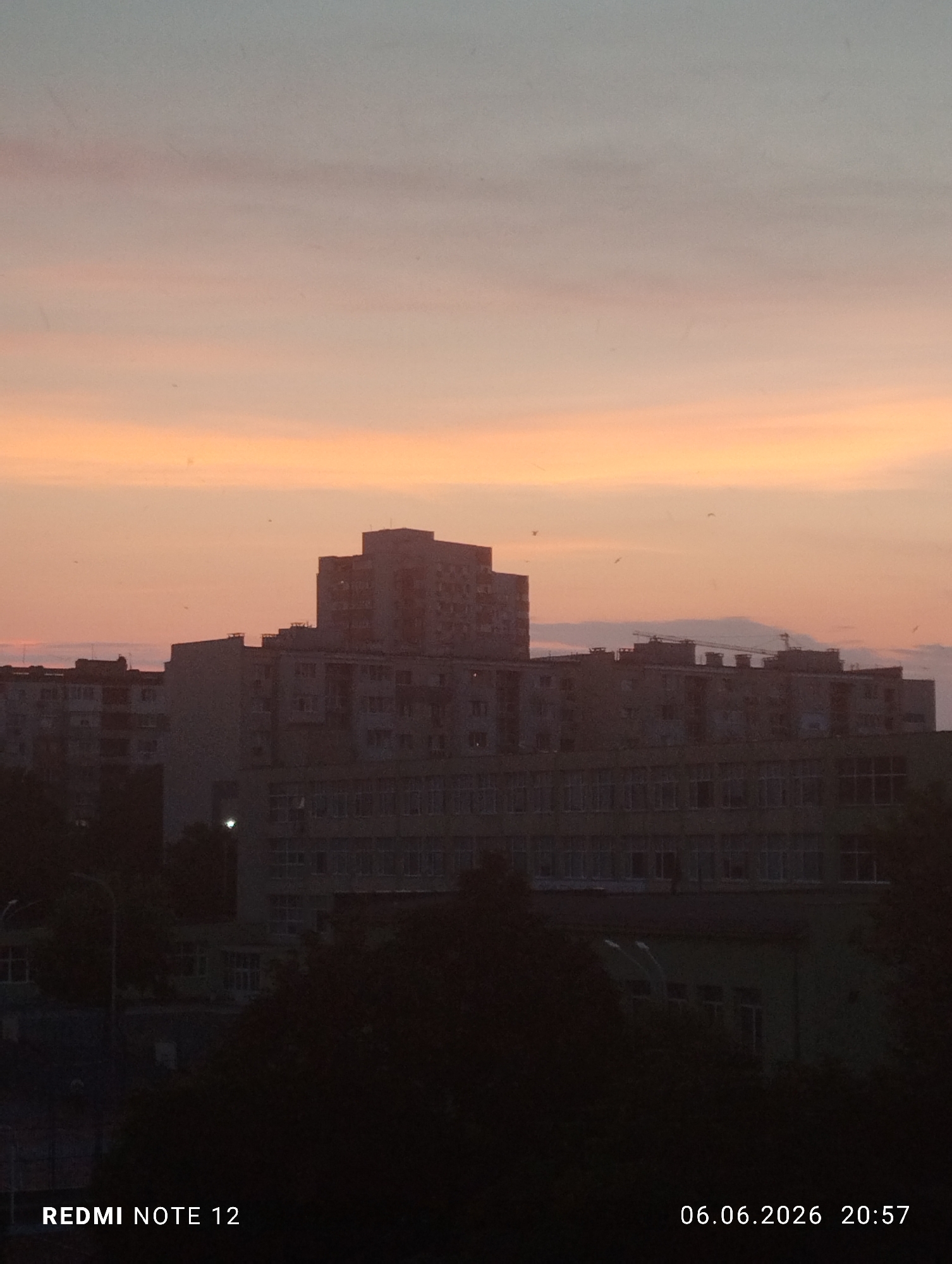 The image depicts an urban landscape at sunset, located in the city of Burgas, Bulgaria, as captured on June 6, 2026, at 20:57 by a Redmi Note 12 device. The upper sky displays a gradient from light blue-grey to subtle pinkish-orange, transitioning into a broad band of orange and pink clouds at the mid-horizon. The lower sky near the horizon is intensely orange. Scattered small dark specks, indicative of birds, are visible against the lighter portions of the sky.
The city skyline is presented in silhouette against the bright sky, revealing multiple multi-story buildings. A prominent, taller building with a stepped profile stands centrally, flanked by other residential or commercial structures of varying heights. On the rooftop of a mid-ground building to the right, a visible structure resembles a crane or construction equipment. The buildings' facades display multiple window arrays, though specific architectural details are obscured by the low light conditions. The immediate foreground is dominated by the dark silhouettes of trees and foliage. A single bright, point-source light, possibly a street lamp, is visible in the lower-left midground. No human figures or direct interactions are discernible within the scene.