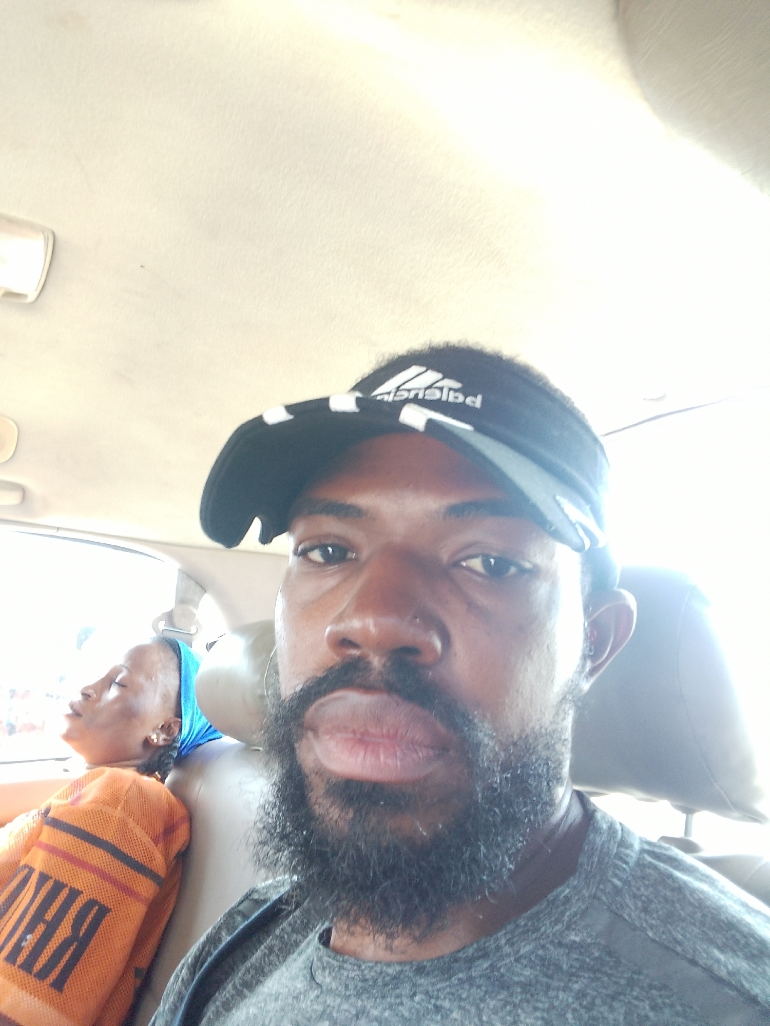 A man with a beard and a black visor hat is in the foreground, taking a selfie in what appears to be a vehicle. He is wearing a gray t-shirt. In the background, a woman with a blue headscarf and an orange and black striped garment is asleep. The interior of the car is visible, including the ceiling and seats. The lighting suggests it is daytime. This scene is occurring within the vicinity of Oworonshoki, Nigeria.