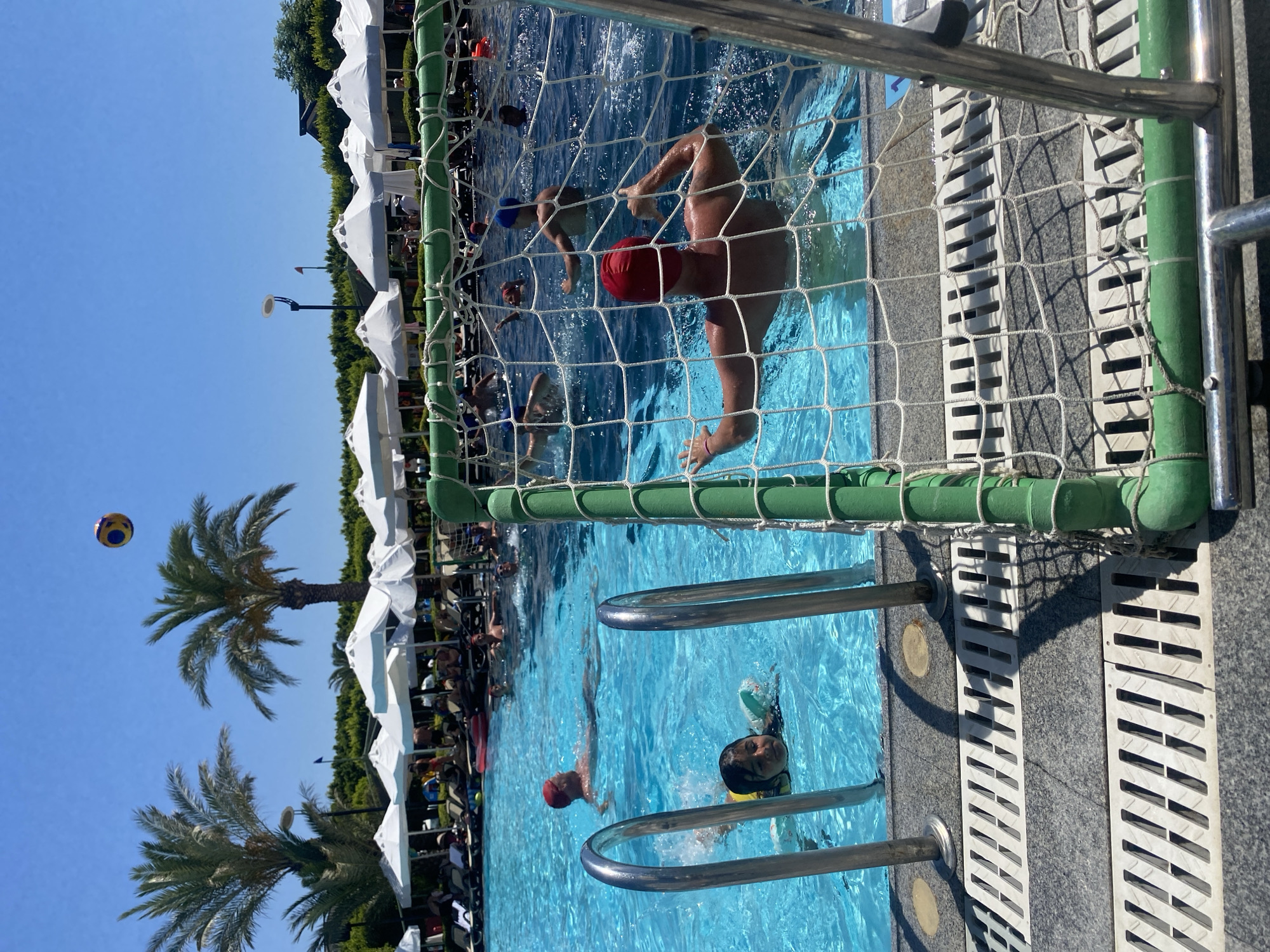 In Aksu, Turkey, a daytime scene unfolds at a resort pool where a water polo game is in progress. The pool is a vibrant blue, with several individuals engaged in the sport. A small child wearing a yellow flotation device swims in the foreground, near a set of stainless-steel pool ladders. In the mid-ground, a man in a red cap is swimming, seemingly participating in the game. Further back, other players are visible, some with blue caps, amidst the splashing water.
The background features a lush green landscape with palm trees and numerous white umbrellas providing shade for lounge chairs along the pool's edge. The sky is a clear, bright blue, indicating fair weather. A water polo goal, with its distinctive green frame and white net, is positioned in the foreground, partially obscuring the view of the game. The net shows signs of use, with some sagging and minor imperfections. The water is clear and reflects the bright sunlight. The overall atmosphere suggests a recreational activity taking place at a holiday destination.