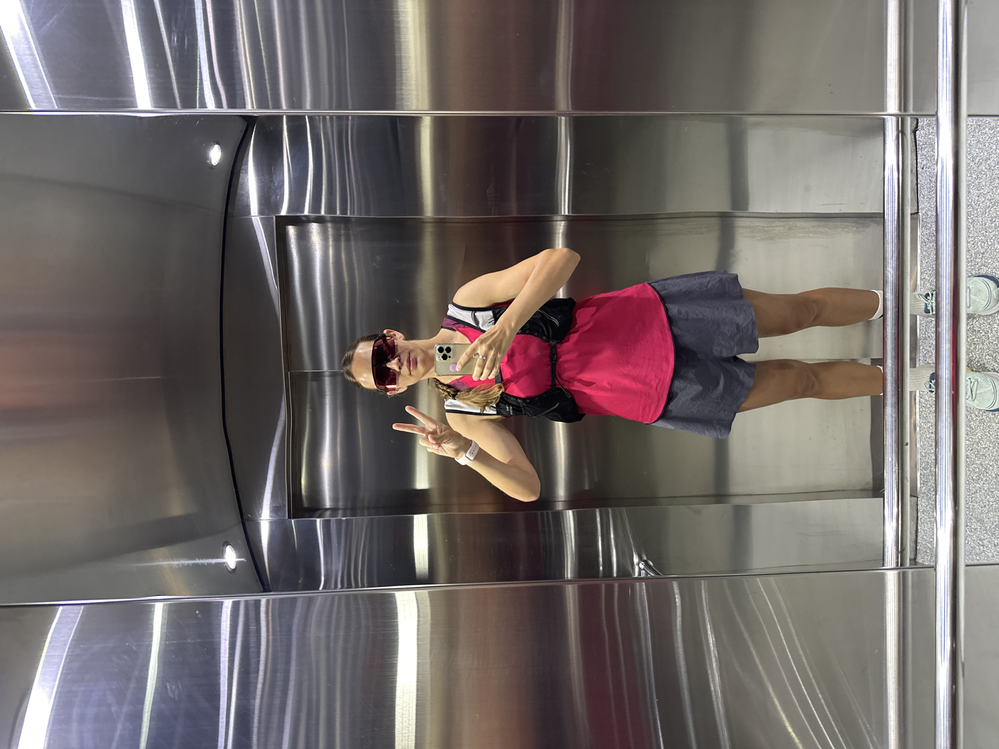 A person stands inside what appears to be an elevator, taking a selfie. The individual has a pink tank top, a dark skirt, and sneakers. They are wearing sunglasses, a smartwatch, and a black fanny pack. The person is holding a mobile phone in their right hand and making a peace sign with their left hand. The elevator has stainless steel panels. A rail and patterned flooring is partially visible. The specific location in Fethiye, Turkey, cannot be determined from the image.