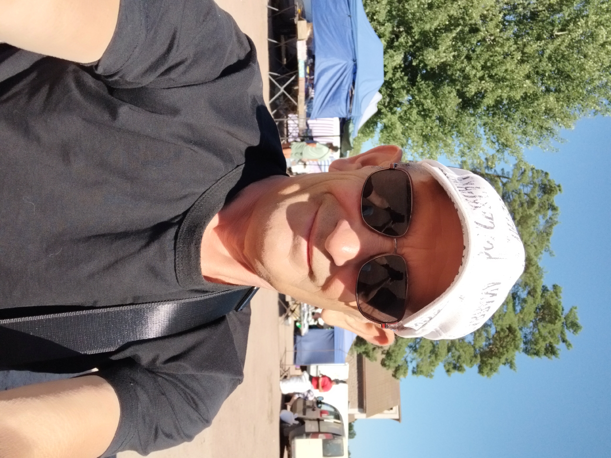 In a medium shot, a man wearing sunglasses and a white baseball cap with a patterned brim smiles at the camera. He is wearing a black t-shirt and a black strap is visible across his chest. The background depicts an outdoor market scene on a sunny day. Two blue tents are set up, with tables laden with merchandise. Further back, a white van and another tent are visible. The ground is a reddish-brown dirt or gravel surface. The man's head and shoulders are the primary focus, with the market serving as a backdrop. The location is not identifiable from the image alone as Irshansk, Ukraine.
