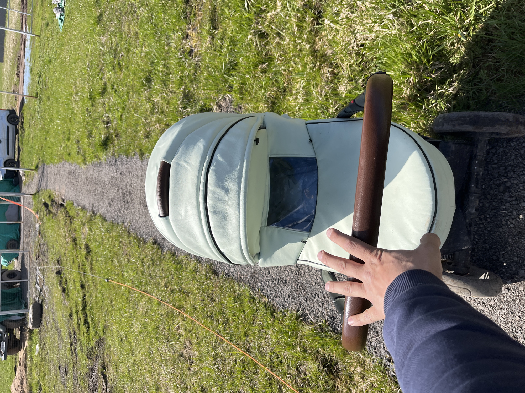 The image shows a person pushing a light green baby stroller on a gravel path. The stroller has a dark brown handle and a window in the hood. The person is wearing a dark blue long-sleeved shirt and their hand is on the stroller handle. The path is surrounded by green grass. In the background, there is a structure with metal framing and a white vehicle with tires. An orange wire or string is visible running along the ground from left to right. The location is Раздолье, Russia.