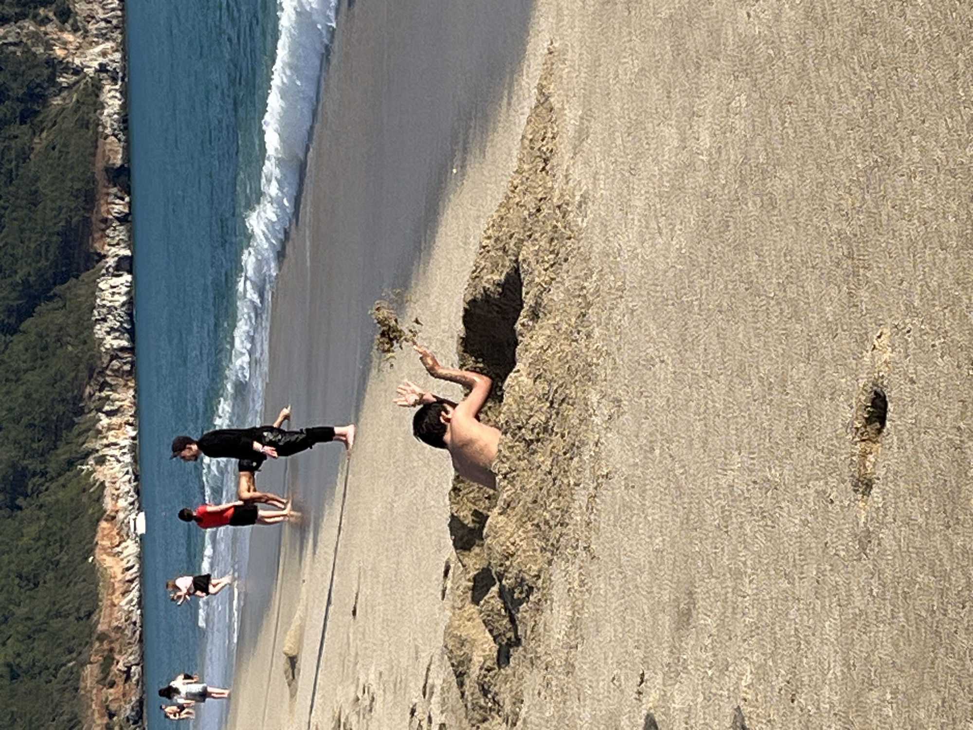 The image shows a beach scene with several people. One person is partially buried in a sand formation on the right side of the frame, with their upper body and arms visible. Other individuals are seen standing and walking near the shoreline on the left, with some appearing to interact or pose. The background includes the sea with small waves, as well as a forested coastline. The scene appears to be located on a beach in Alanya, Turkey, as indicated by the vegetation and coastal features.
