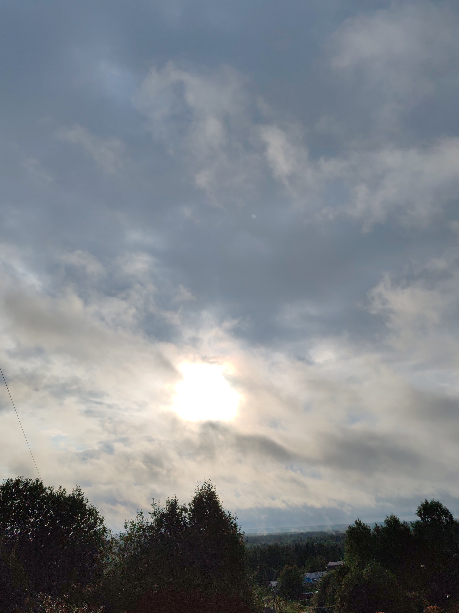The image depicts an outdoor scene, likely taken in a rural or suburban area near Чухлэм, Russia. The foreground is dominated by the dark silhouettes of trees, indicating either early morning or late evening light, or a heavy overcast sky. Above the trees, a partially obscured sun shines brightly through a thick layer of gray and white clouds, creating a diffused and somewhat hazy light. The sky takes up the majority of the frame. In the distance, below the tree line, a landscape of more trees and some hints of structures, possibly houses or buildings, can be seen. A single thin wire, possibly a power line or telephone line, extends diagonally from the top left corner towards the trees. The overall impression is one of a moderately cloudy day in a natural or semi-natural environment.