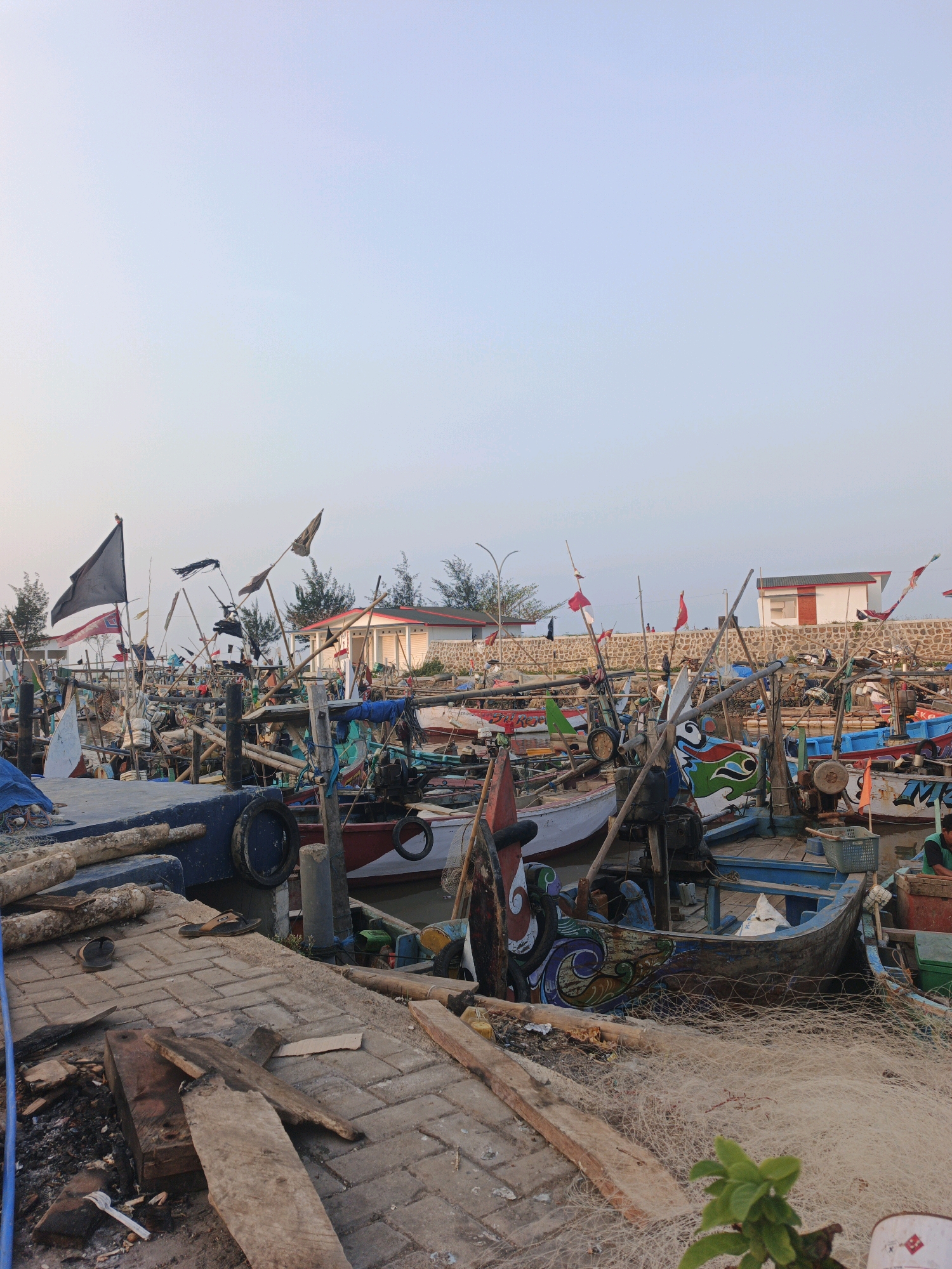 The image depicts a fleet of traditional Indonesian fishing boats docked at a harbor in Bumiharjo, Indonesia. The boats, varying in size and color, are adorned with decorative paintings and flags. Some boats have nets piled on deck, suggesting recent fishing activity. The harbor itself appears to be a mix of concrete and stone structures, with wooden planks and debris scattered along the waterfront. In the background, a few small buildings with red roofs are visible, along with trees and a stone wall. The sky is clear and pale, indicating either early morning or late afternoon. There are no visible people actively engaged in any tasks, but the scene suggests a busy working harbor.