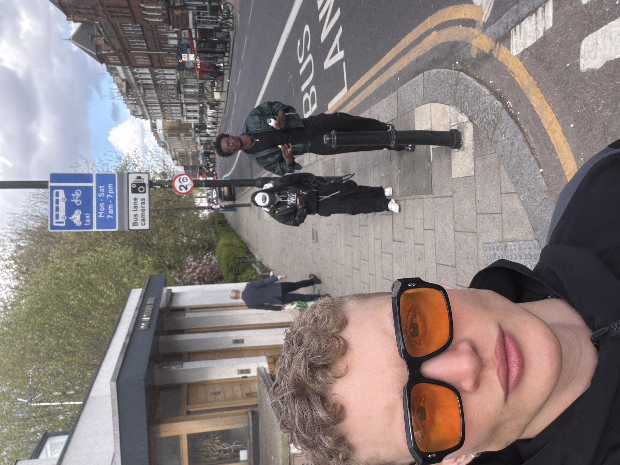 The image is a selfie taken by a young person with curly hair and orange-tinted sunglasses. In the background, two people are standing on the edge of a bus lane on a street in London, United Kingdom. One person is wearing a green jacket and black pants and holding something in their hand, while the other is wearing a black jacket, black pants, and a white hat. There is a signpost indicating a bus lane operating from Monday to Saturday between 7 am and 7 pm, as well as a sign indicating bus lane cameras and a 20 mph speed limit. Buildings line the street in the background.