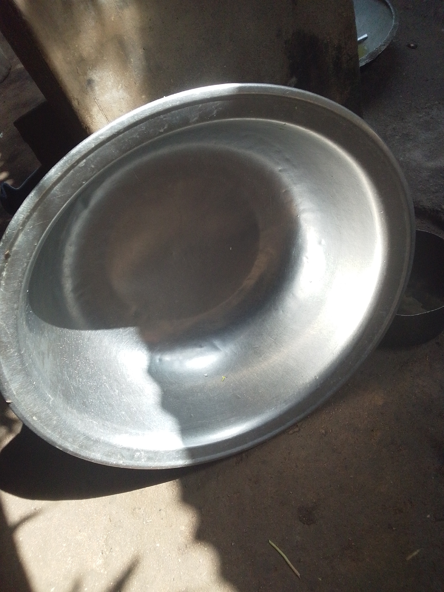 An outdoor, eye-level, close-up shot shows a large, shallow, circular, metallic basin on a rough, dirt surface. The basin is angled towards the viewer from the right, with its rim visible along the bottom and right edges of the frame, and curving upwards towards the top left. The interior of the basin reflects light brightly, creating a gradient of shadows and highlights. The surrounding environment is dimly lit, with shadows cast from an unseen object above and to the left, creating a dappled pattern on the basin and the ground. To the right and slightly behind the basin, a dark, cylindrical object is partially visible, its rim also appearing metallic. The background shows a textured, dark surface, likely the ground, with some debris. The overall impression is of a utilitarian setting, possibly in Jalingo, Nigeria, where everyday objects are photographed in natural light.