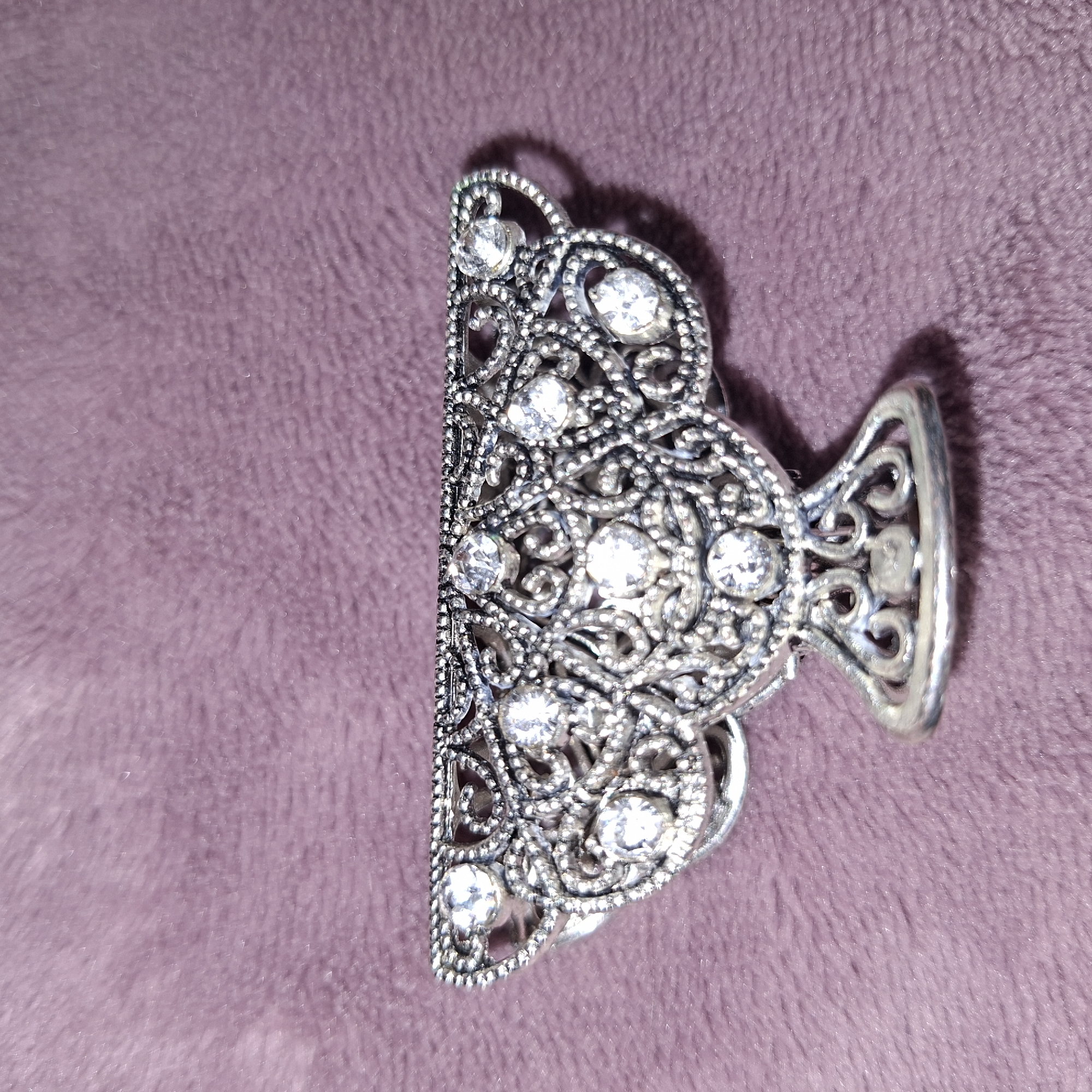 The image displays a silver hair claw adorned with small clear rhinestones. The hair claw has an intricate, filigree design with swirling patterns and a beaded edge. It is positioned on a textured, purple fabric surface, which appears soft and possibly velvet-like. The lighting casts a subtle shadow from the hair claw onto the fabric, indicating the light source is coming from the right. The image is a close-up shot, focusing primarily on the hair claw and the immediate surrounding fabric. There is no visible text, people, or activity depicted in the image. The setting appears to be indoors, given the controlled lighting and fabric backdrop. The specific location of Mingachevir, Azerbaijan, is provided as context but is not discernible from the visual elements of the photograph itself. The overall impression is one of an accessory detail shot, highlighting the craftsmanship and sparkle of the hair claw.