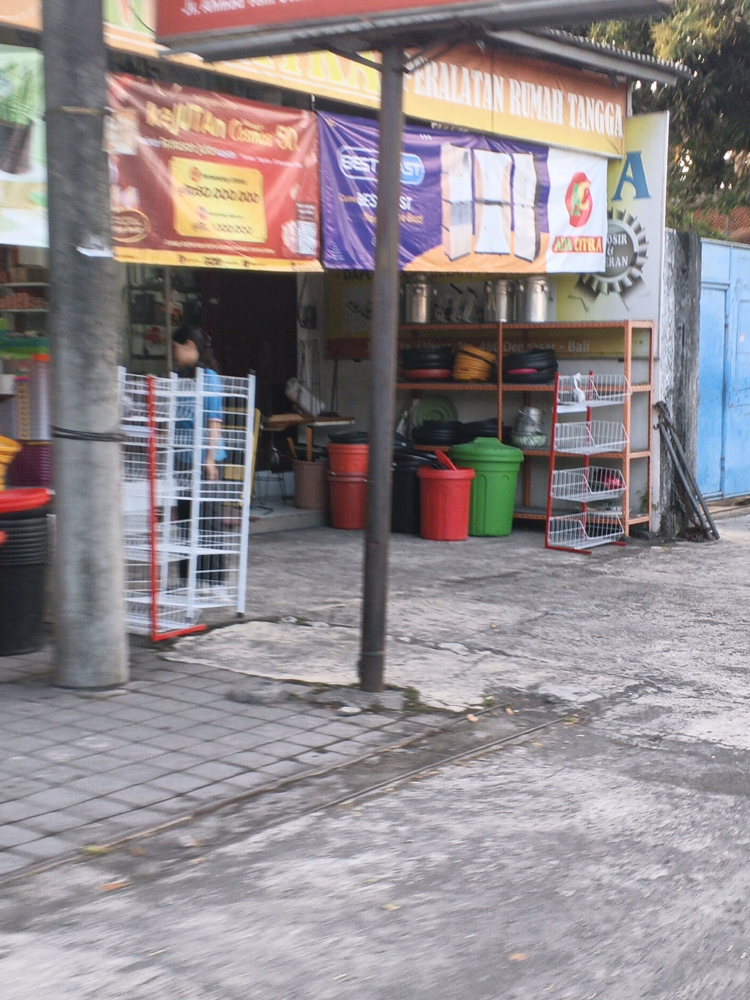 This is a full-shot, eye-level, exterior, horizontal, daytime, over-the-shoulder shot of a street scene in Denpasar, Indonesia. The image captures a storefront with banners and displays of household goods. A large, cylindrical, gray concrete pillar is prominent in the foreground on the left. Behind it, a white metal display rack with red trim holds various items, and a person in a blue shirt is partially visible inside.
To the right of the pillar, a red banner with yellow lettering reads "KEJUTAN COSMETIC 50." Further to the right, a purple banner with white text is visible. Above these, an orange banner with white lettering states "PERALATAN RUMAH TANGGA."
A retail display unit with orange shelving holds various items, including what appear to be buckets and baskets. In front of this unit, there are two large plastic trash cans, one green and one red. To the far right, a blue door is visible, and a set of dark metal poles lean against a wall. The ground is paved with gray square tiles, and a wet, asphalt-like surface with some debris is present. The overall impression is a commercial area with a focus on household and cosmetic products.