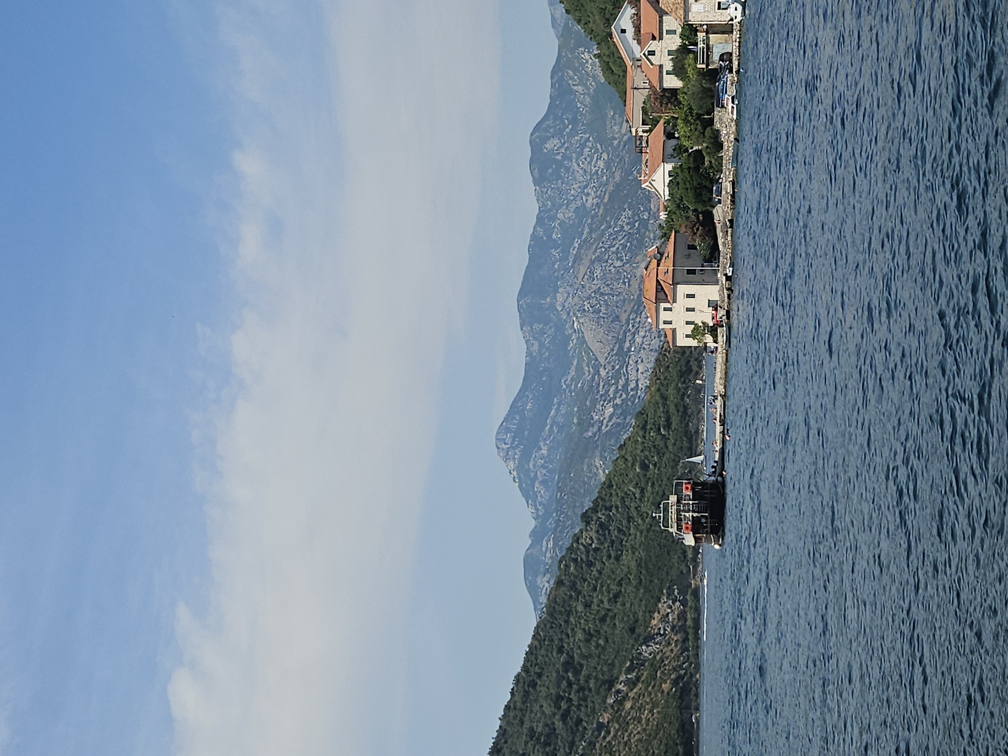 The image captures a serene daytime scene in Lepetane, Montenegro, featuring a waterfront view. In the foreground, the deep blue water of the bay is visible, with gentle ripples across its surface. On the left side of the image, a ferry is docked near a lush, green hillside. The ferry is dark-colored with a white upper deck and appears to be stationary.
In the mid-ground, a small coastal town with several buildings is situated along the water's edge. These buildings are characterized by their terracotta-tiled roofs and light-colored facades. Some of the buildings are residential homes, while others may be part of the town's infrastructure. A paved walkway or road runs along the waterfront in front of the buildings, with parked cars visible.
The background is dominated by a majestic mountain range, its peaks and slopes a mix of rocky grey and green vegetation. The sky above is a clear blue, with scattered white clouds adding depth and texture. The overall impression is one of a peaceful coastal settlement nestled between the mountains and the sea.