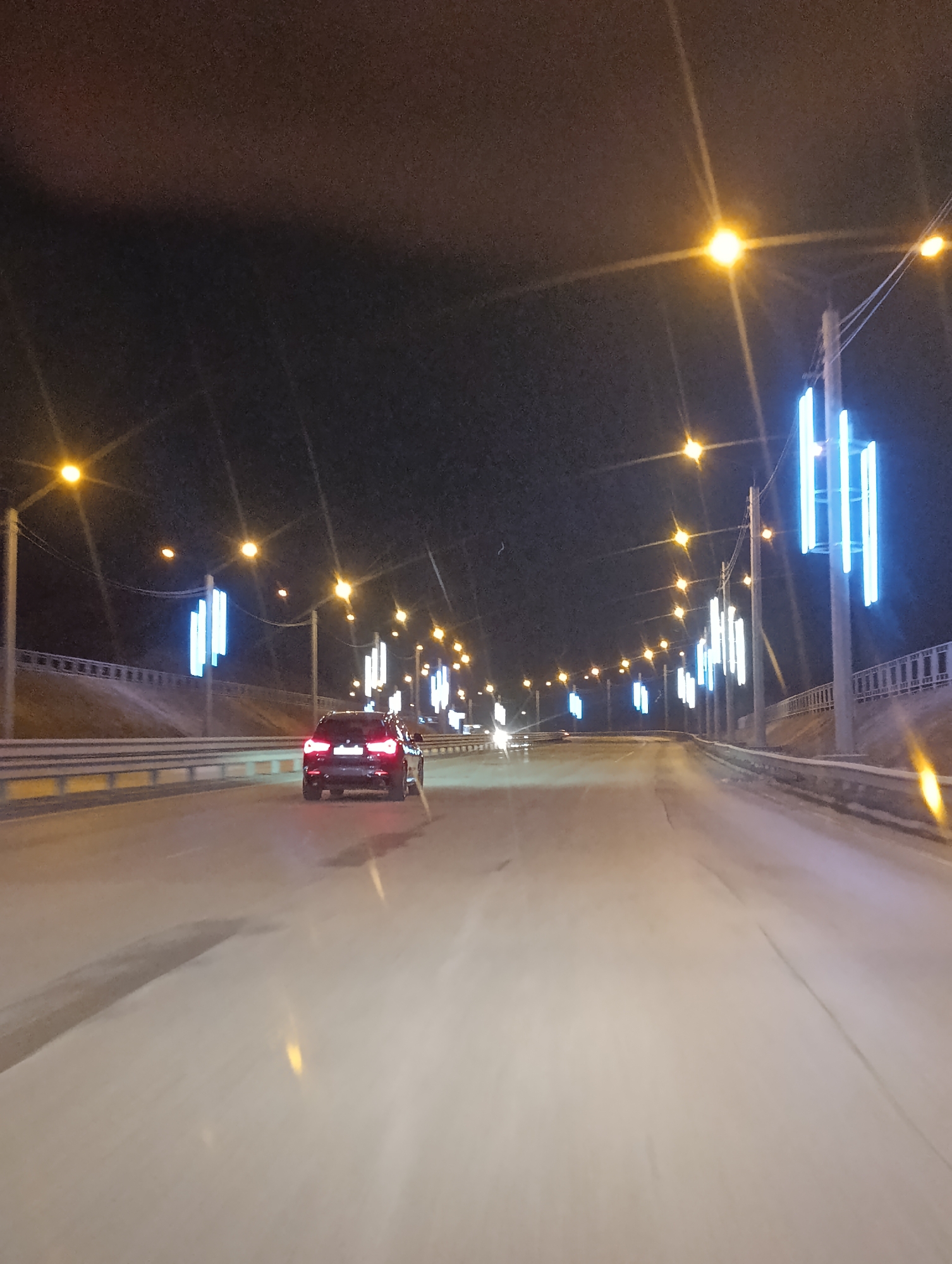 The image depicts a nighttime scene on a multi-lane road in Krasnoyarsk, Russia. A dark-colored car with illuminated rear lights is traveling away from the viewer. Rows of streetlights line the road, some with additional illuminated blue decorative elements. A concrete barrier is visible on either side of the road. The road surface appears to be wet or icy. The overall environment is dark, illuminated primarily by artificial lights.