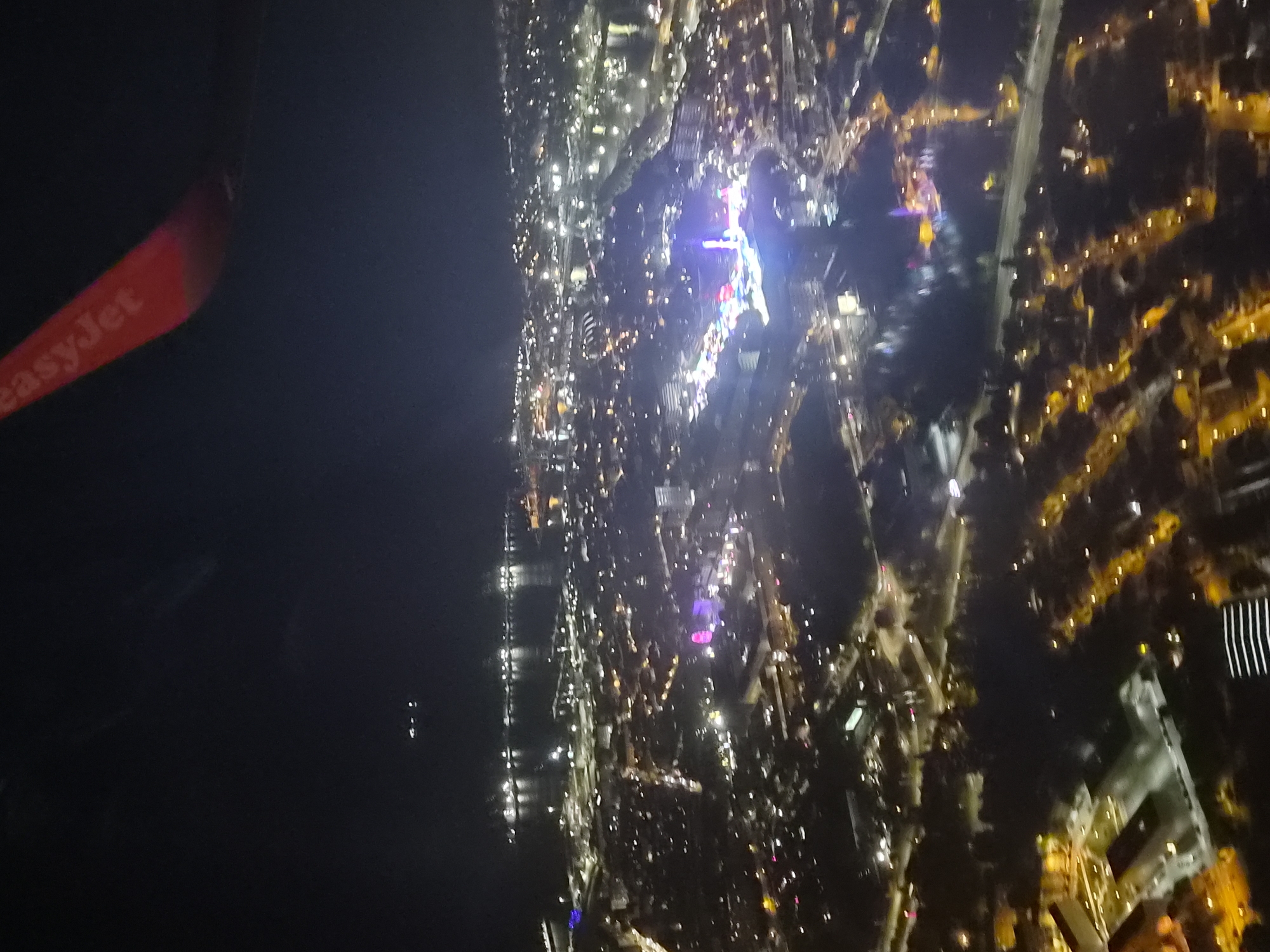 A nighttime aerial view from an EasyJet plane, with a city landscape. The city is illuminated by numerous lights, predominantly yellow and white. The angle indicates that the city is along a coast with water on the left side of the view. The city has many streets and buildings and a few areas of distinct purple and blue lighting. The image is blurry, indicating movement of the plane. The location is Matosinhos, Portugal.