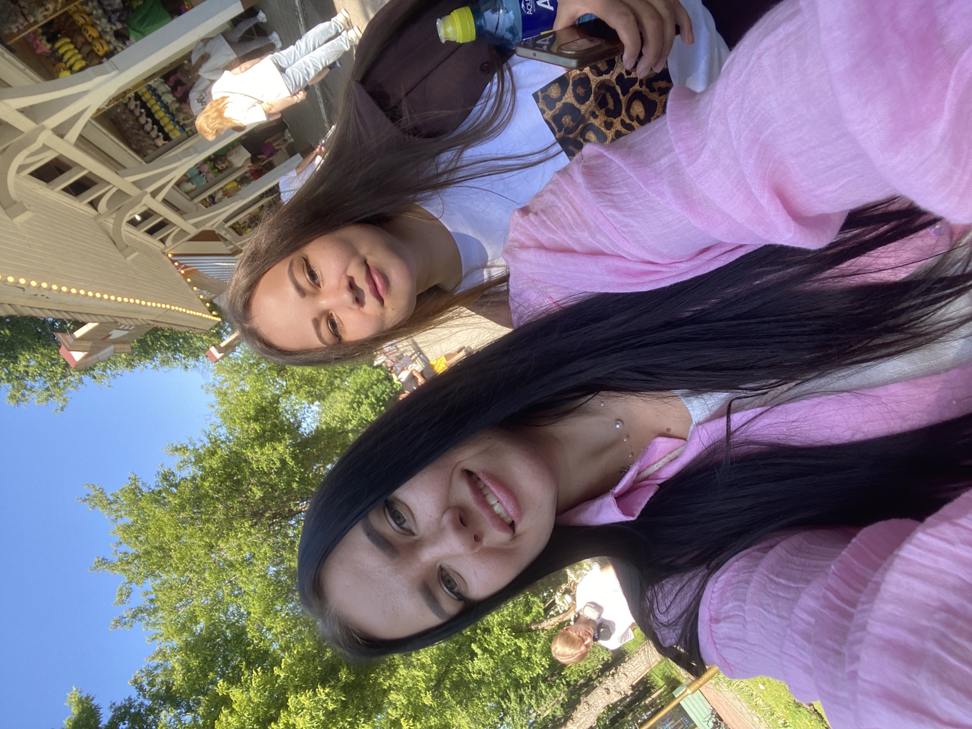 In a selfie taken outdoors on a sunny day, two young women are positioned in the foreground. The woman on the left, with long dark hair and wearing a pink shirt, smiles at the camera. The woman on the right, with long brown hair and wearing a dark jacket over a white shirt, also looks towards the camera with a slight smile. A bottle of water and a leopard-print clutch are visible in her hands. Behind them, a white building with decorative lights is visible, suggesting an amusement park or a similar recreational area. Trees and a blue sky are in the background. The overall scene suggests a casual outing in a public, likely urban, setting.