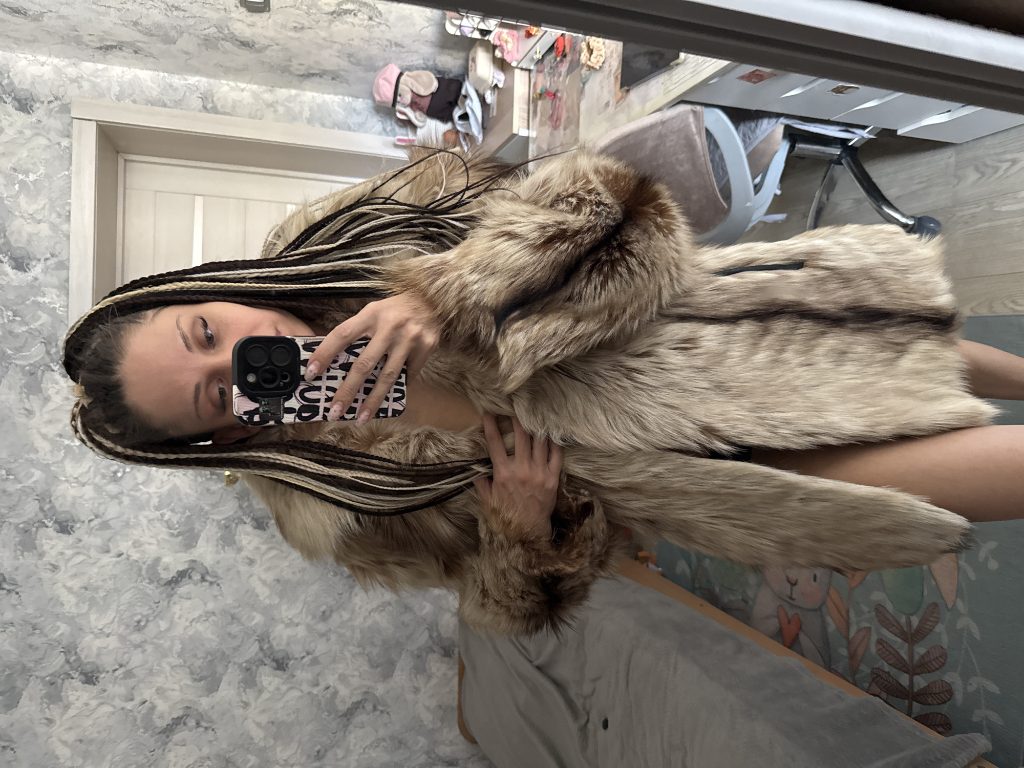 A woman with brown and blonde braids is taking a selfie in a mirror. She is wearing a fur coat. The background includes a grey wall with a white door frame, a clothes pile, a chair, and a bed with a patterned blanket. This location is not identifiable as Bor, Russia based on visual information.