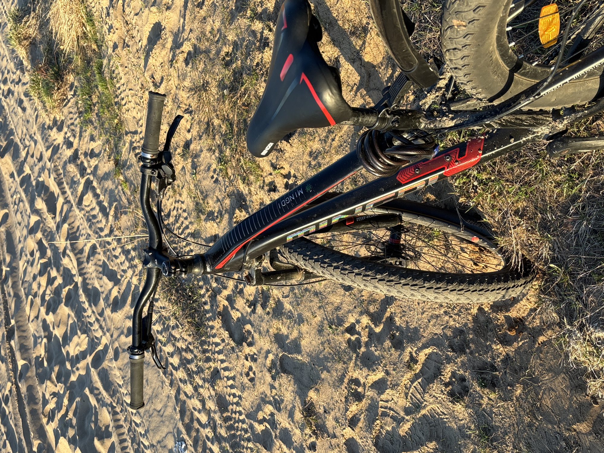 The image shows a black and red mountain bike lying on its side in a sandy or dirt-covered area with sparse vegetation. The bike has knobby tires, suggesting it's designed for off-road use. The saddle is black with red accents. The frame displays the brand name "MINGDI". The setting is outdoor, with evidence of tire tracks in the sandy ground. The location is Chaikovsky, Russia.