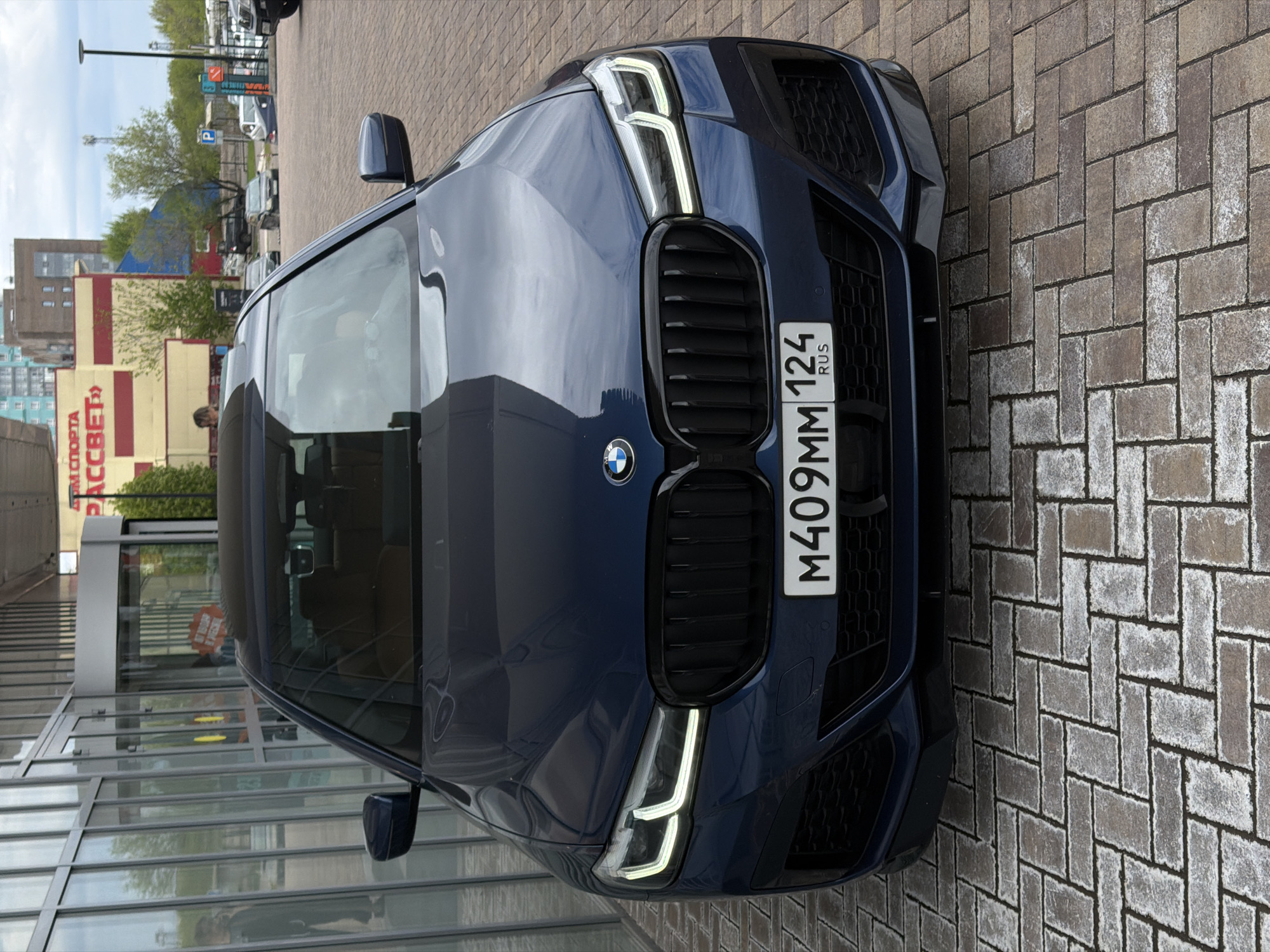 A blue BMW is parked on a brick-paved surface in Krasnoyarsk, Russia. The car's front is facing the viewer with the license plate reading "M409MM 124 RUS." The background includes a multi-story building with a sign reading "PACCCBET" (Рассвет) which is visible behind the car. There is also a glass-walled building visible on the left. The environment appears to be urban and the weather seems to be overcast or partly cloudy. Other vehicles and trees can be seen further in the background.