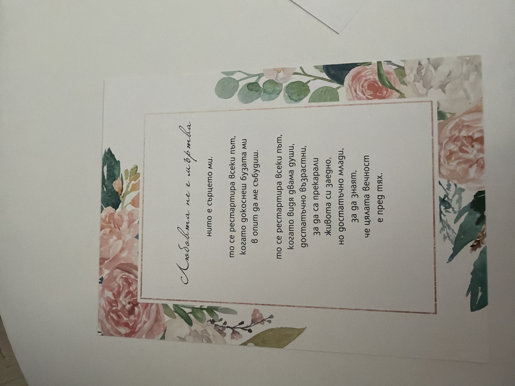 The image shows a card with floral decorations and text in Bulgarian. The text is written in a cursive font. The card is positioned on a flat, white surface, possibly a table or a desk. The background is out of focus and appears to be a wall. There are no people or specific locations identifiable in the image. The scene is static and appears to be a still life of the card.